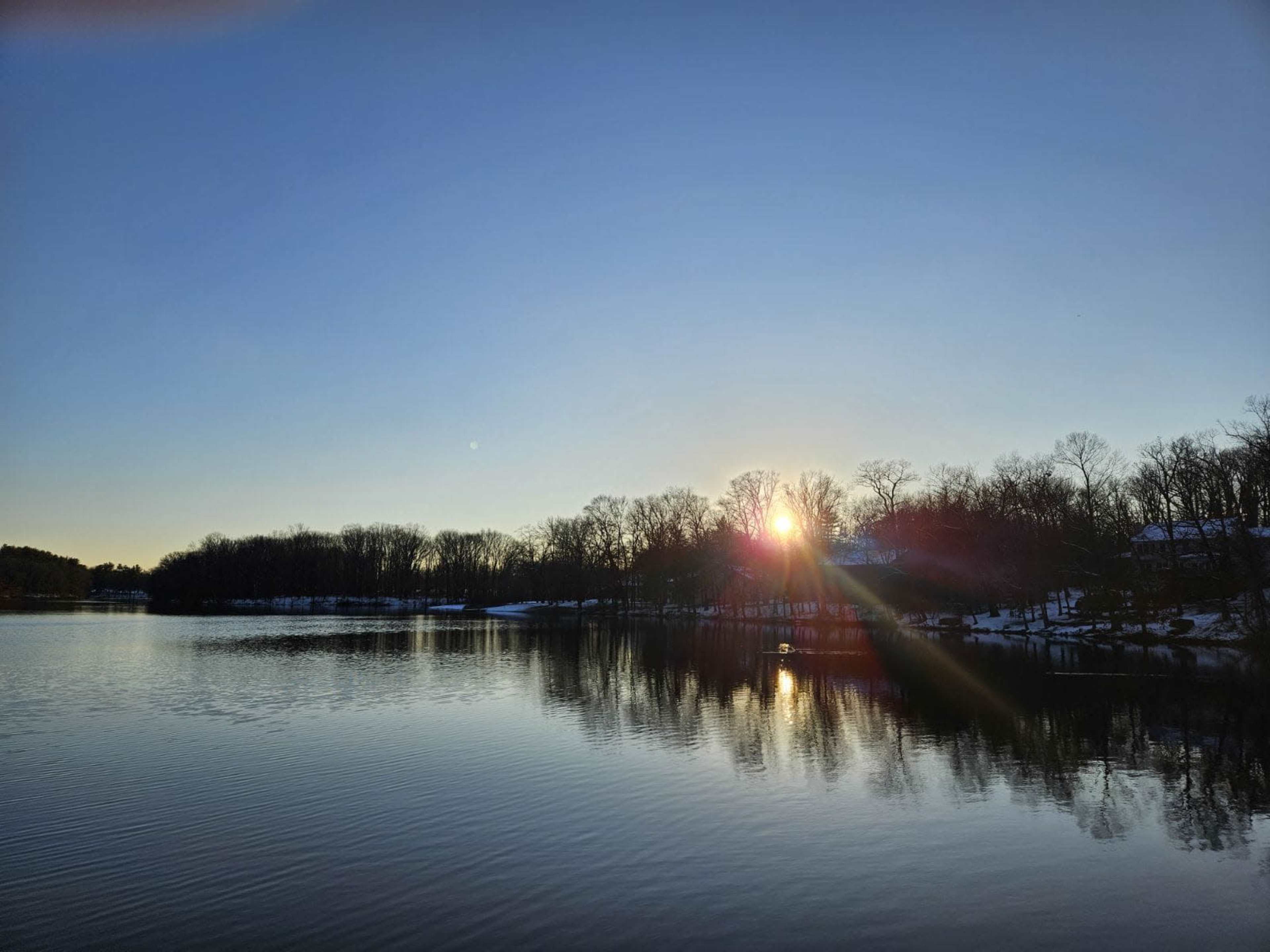 The sun rises over a calm lake, reflecting the trees and clear sky.