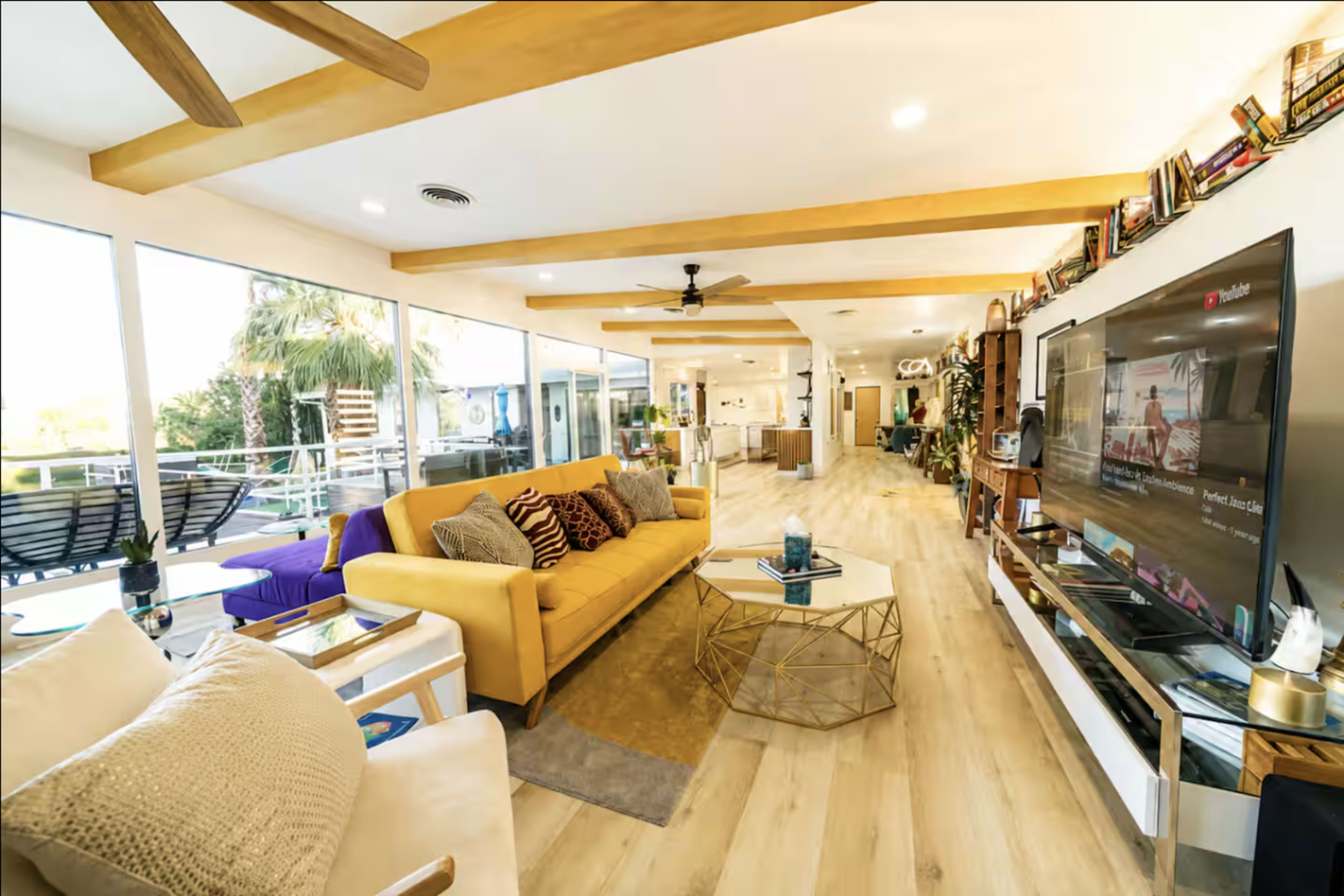 The image shows a modern living room with a yellow sofa, a coffee table, and a large TV, surrounded by large windows that let in natural light.