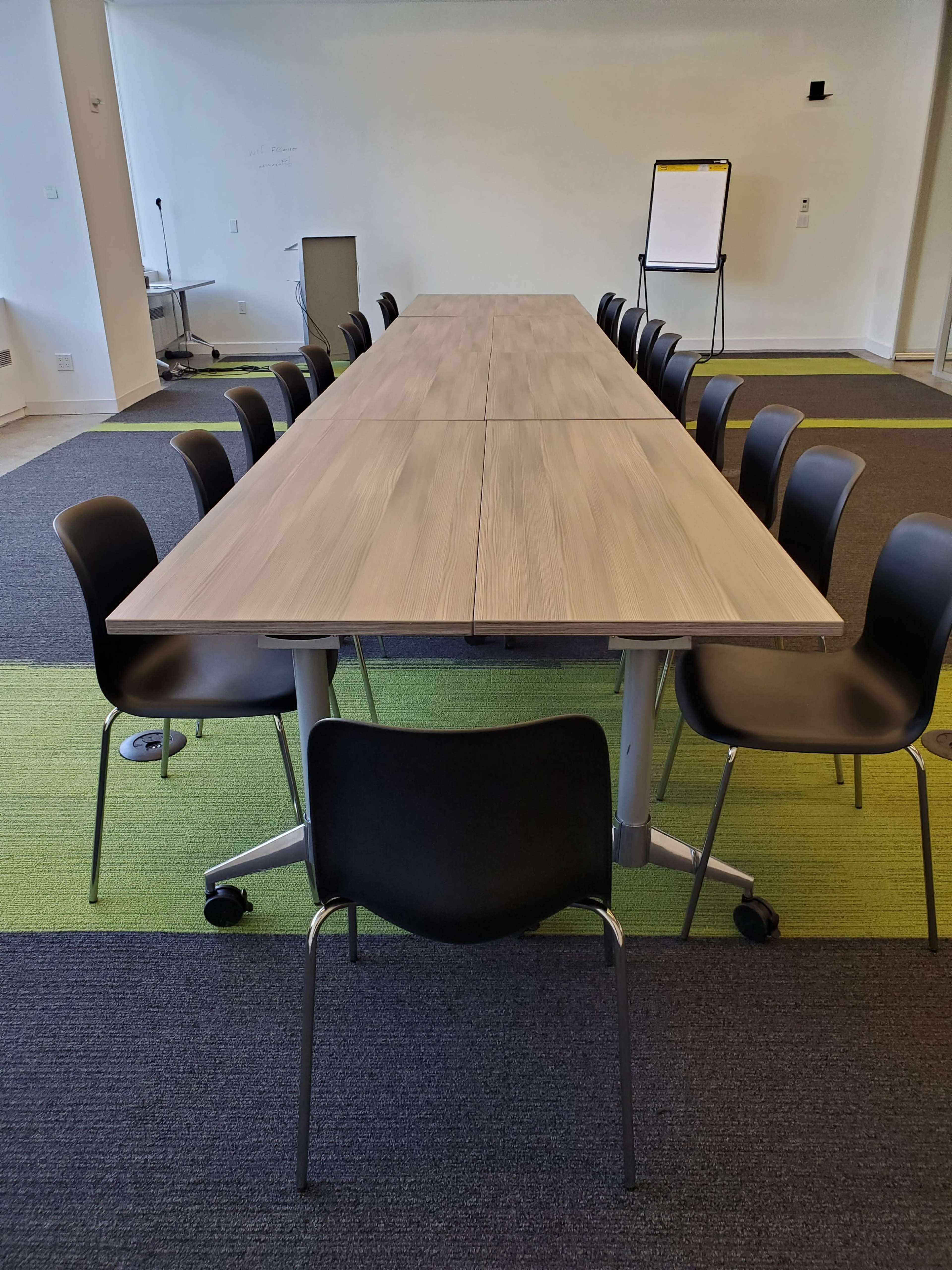 A long, rectangular table with rolling black chairs is set in a conference room with green and gray carpeting.