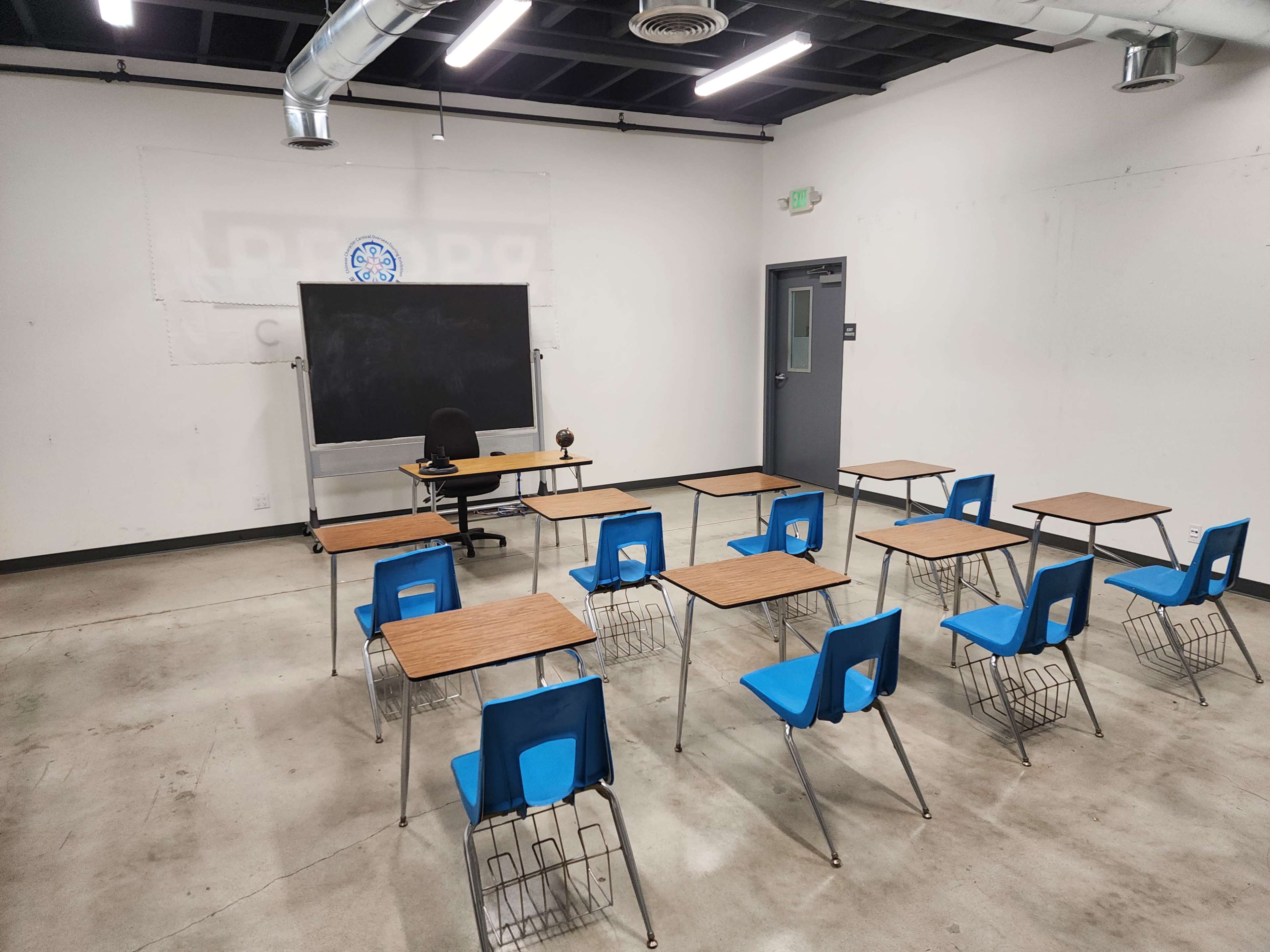 A classroom is arranged with blue chairs and wooden desks facing a chalkboard and an empty teacher's desk.