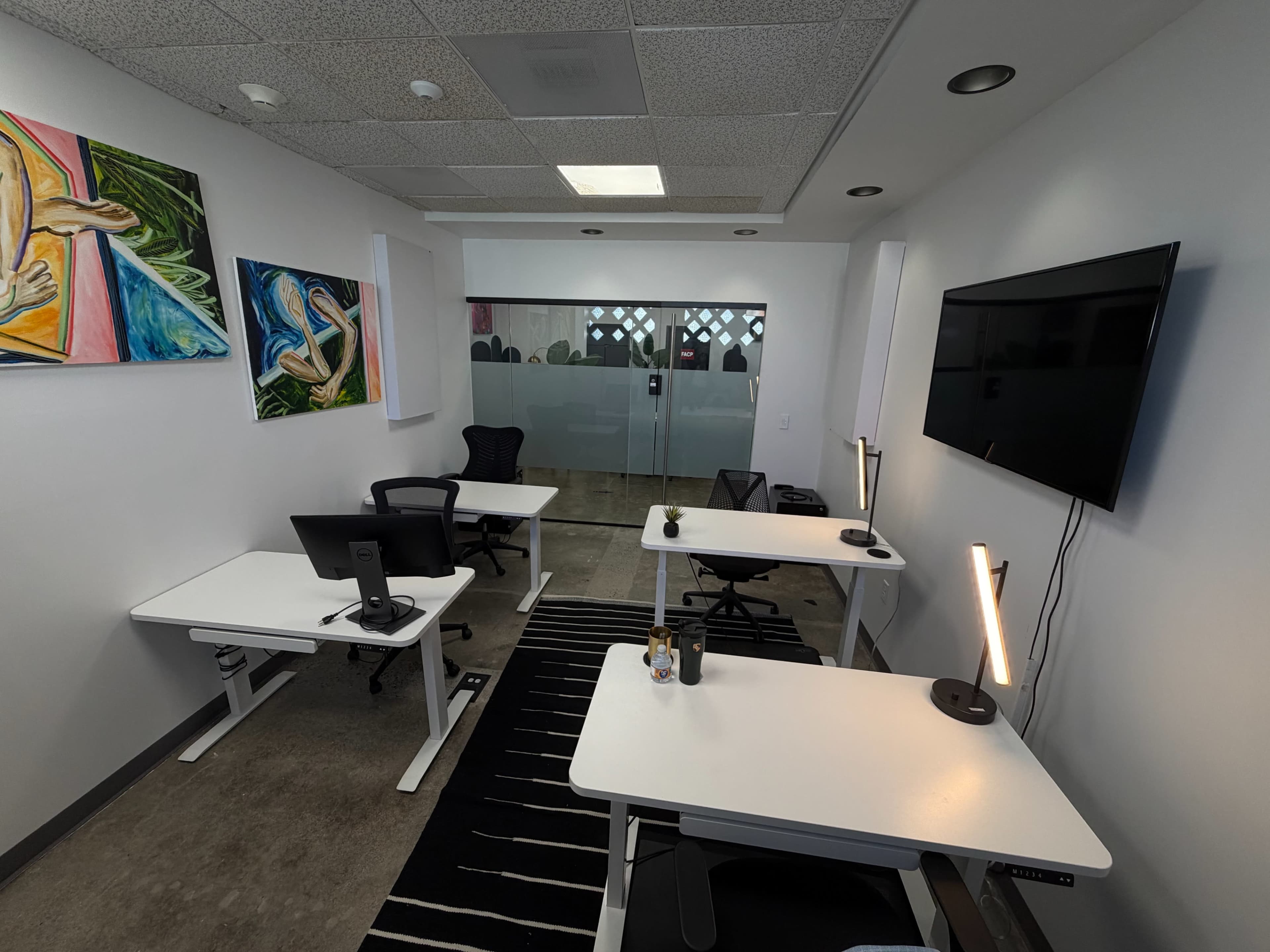 The image shows a modern office space with two desks, each equipped with monitors, a television on the wall, and colorful artwork displayed.