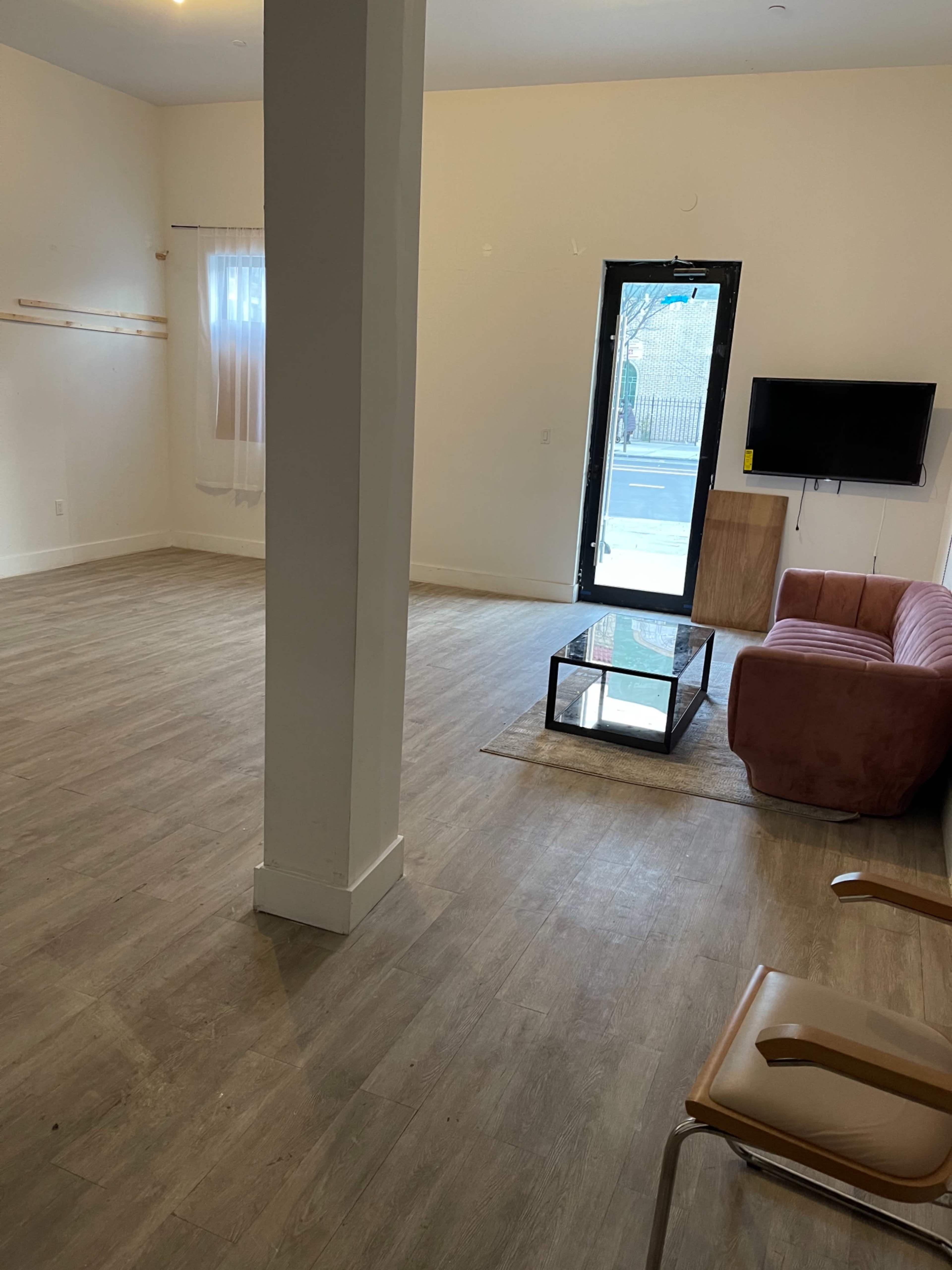 The image shows a spacious, empty room with a wooden floor, a pink sofa, a glass coffee table, a television mounted on the wall, and a large window with a view outside.