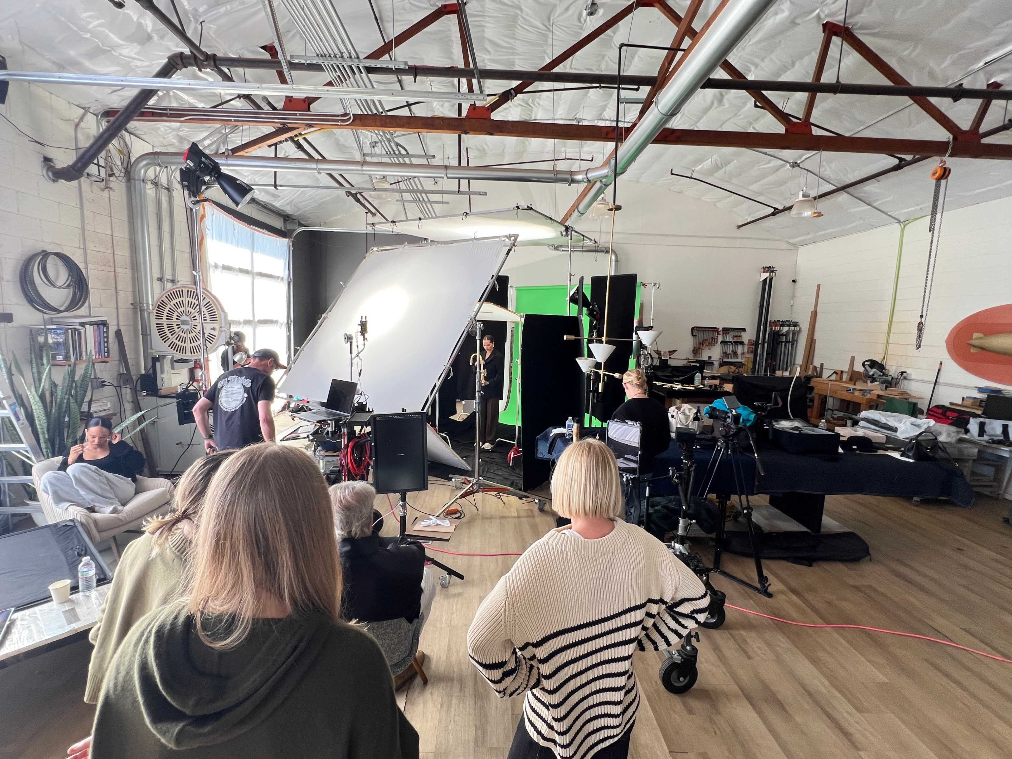 A group of people observes a filming setup in a studio, featuring lights, cameras, and a large backdrop.