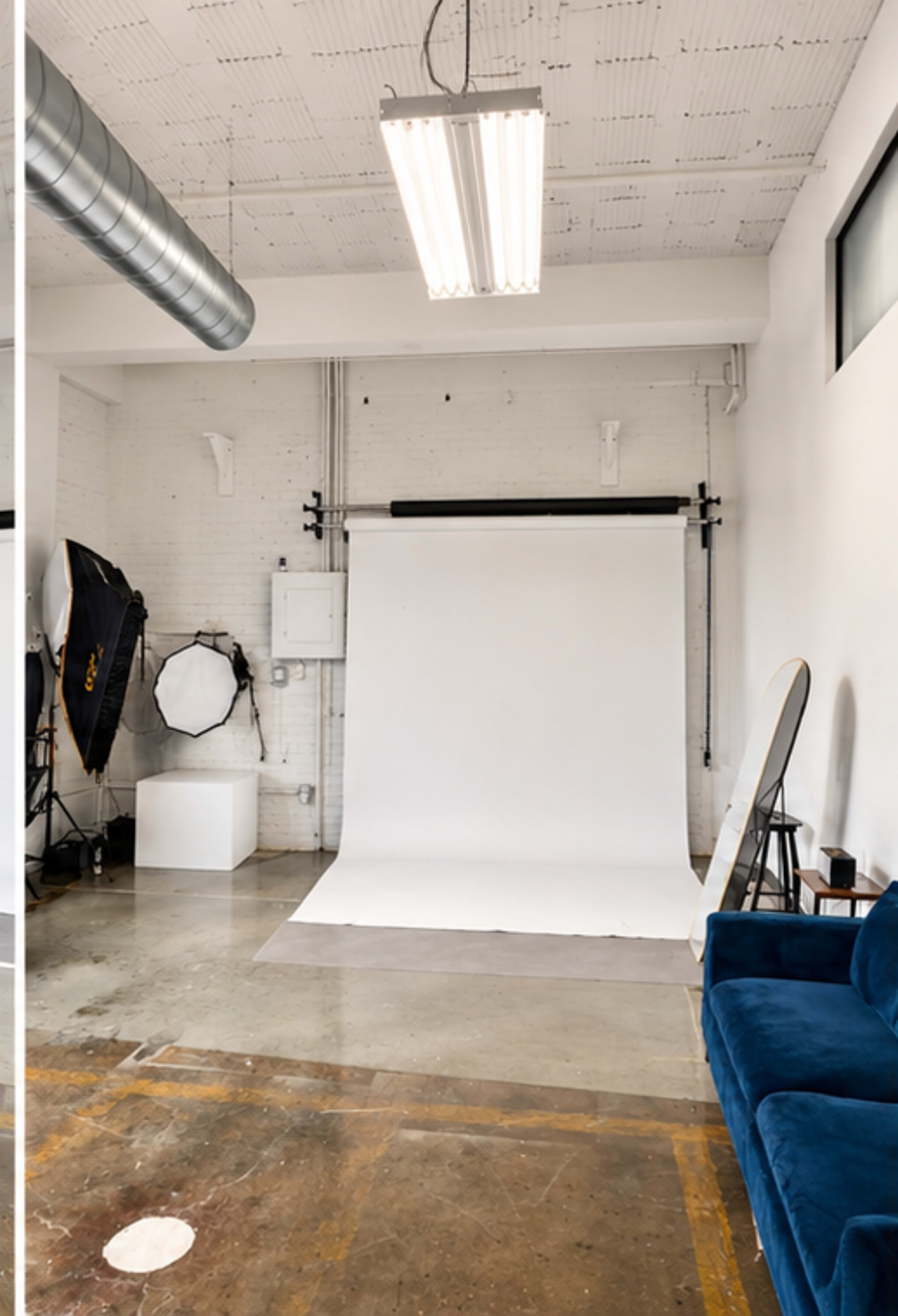 The image shows a minimalist photography studio with a white backdrop, a blue couch, and various lighting equipment.