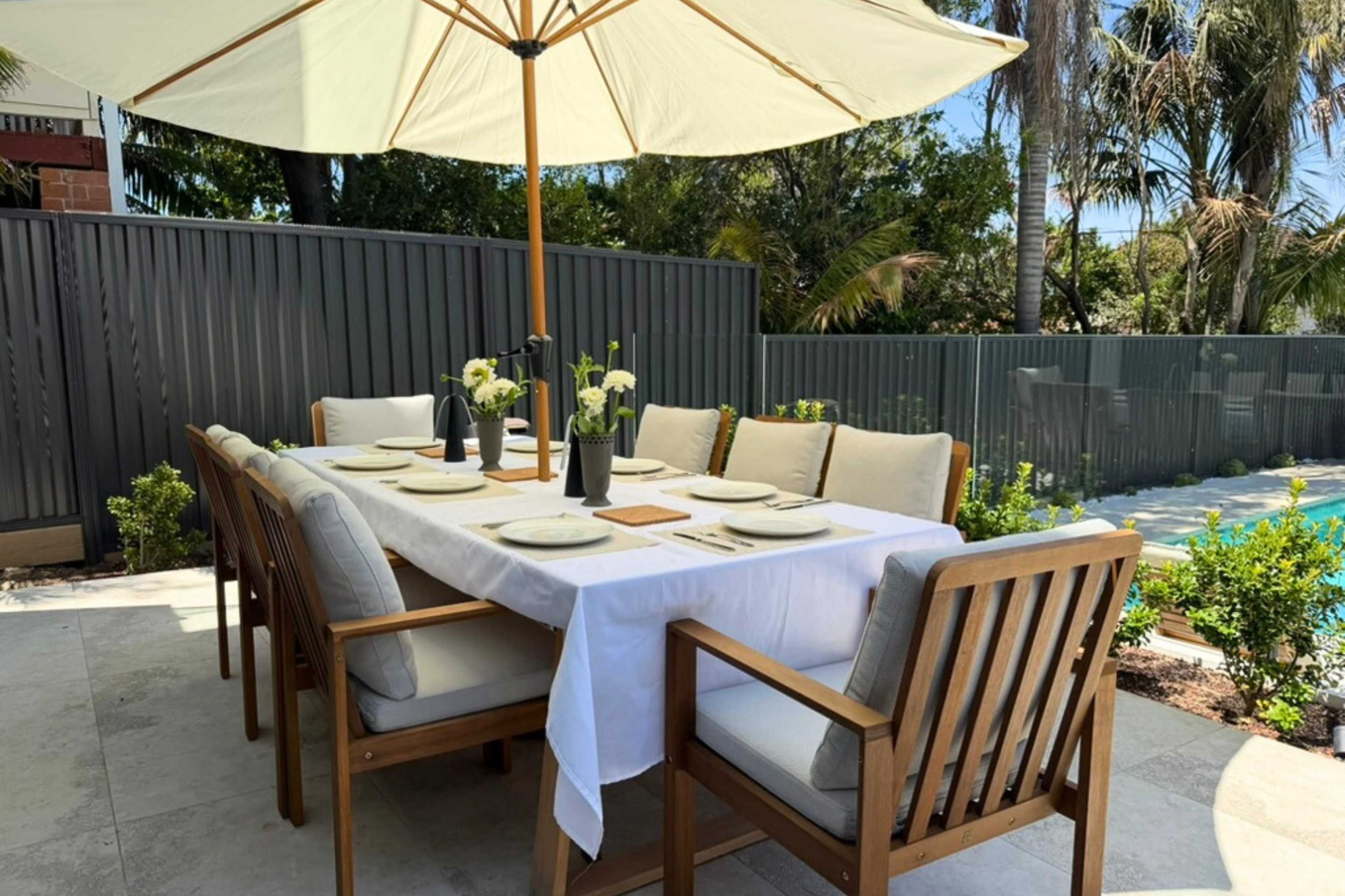 A wooden dining table with white tableware is set under a large umbrella beside a pool, surrounded by green plants and a gray privacy fence.