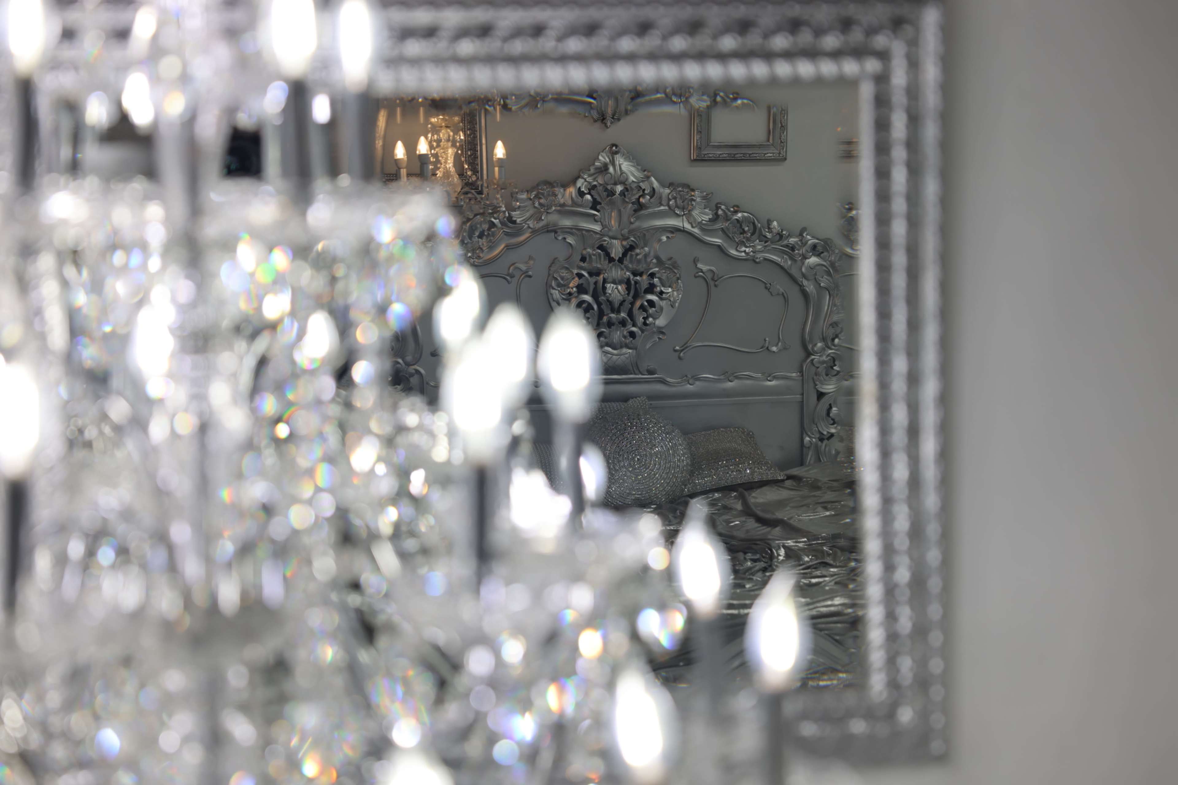 The image shows a reflection of an ornate silver bed in a decorative mirror, with a chandelier in the foreground.