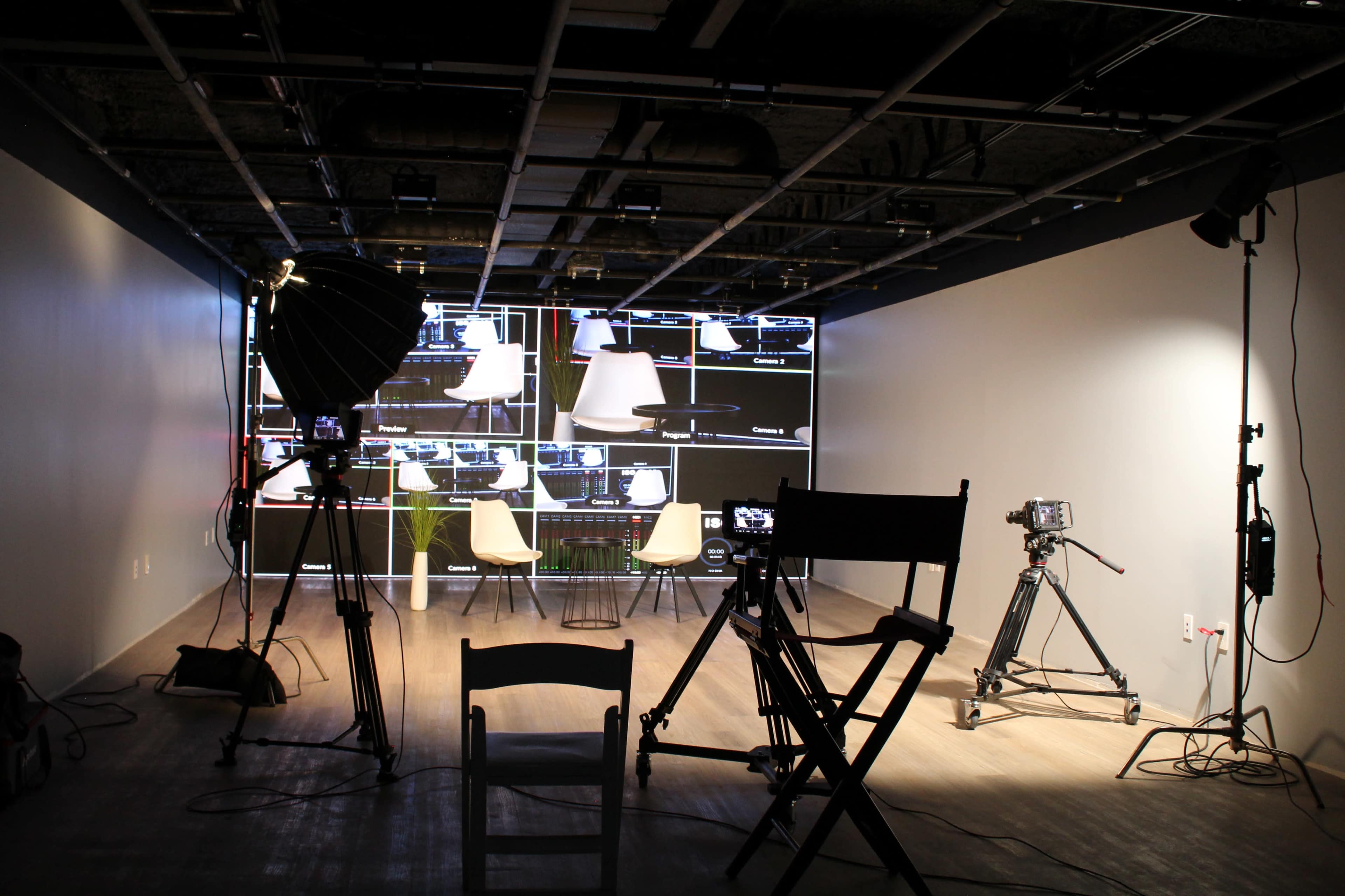 A video recording studio is set up with two chairs facing a large screen displaying multiple screens, along with various cameras and lighting equipment.