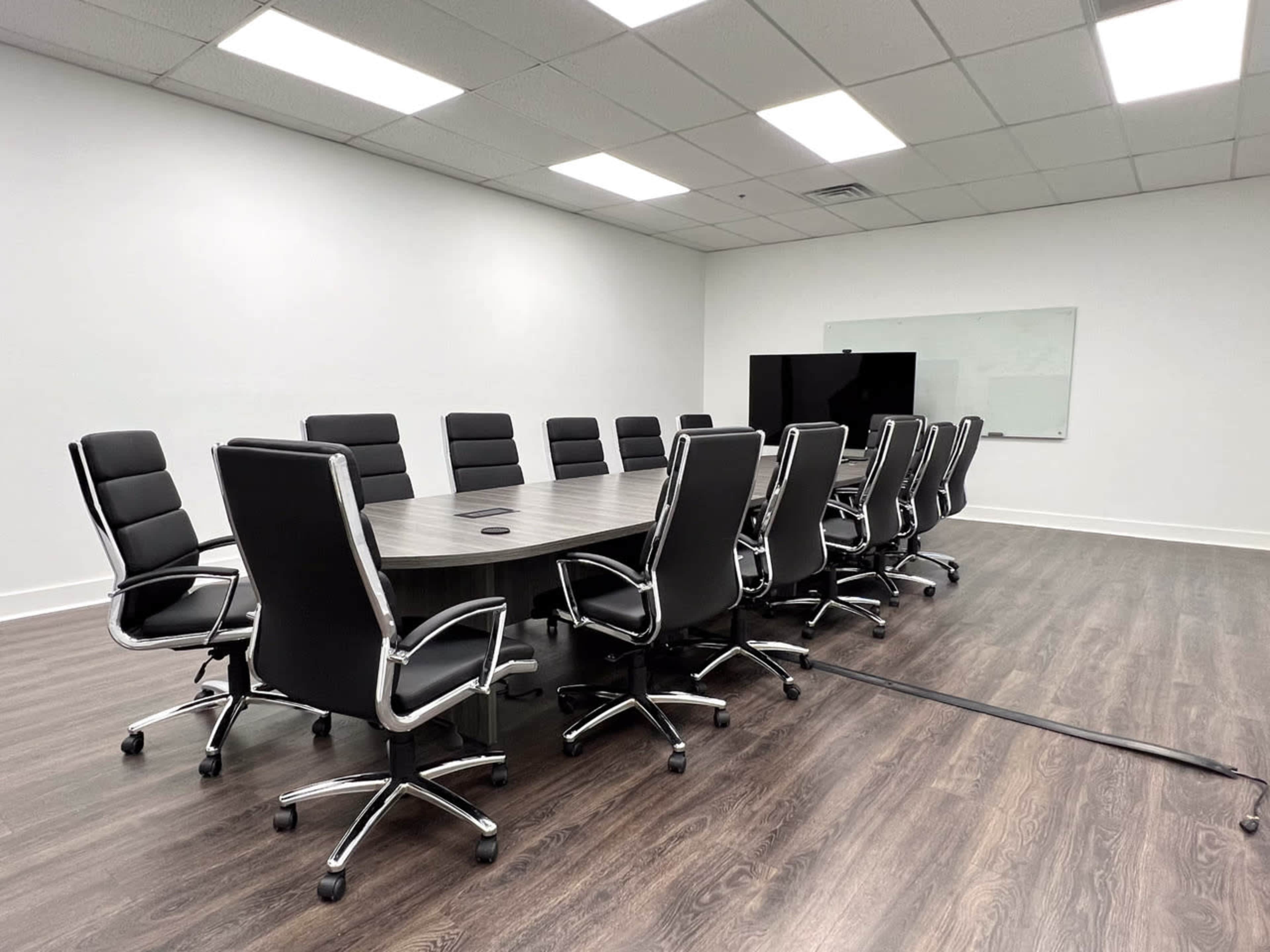 A modern conference room features a large round table surrounded by twelve black ergonomic chairs, with a whiteboard and a television screen on one wall.