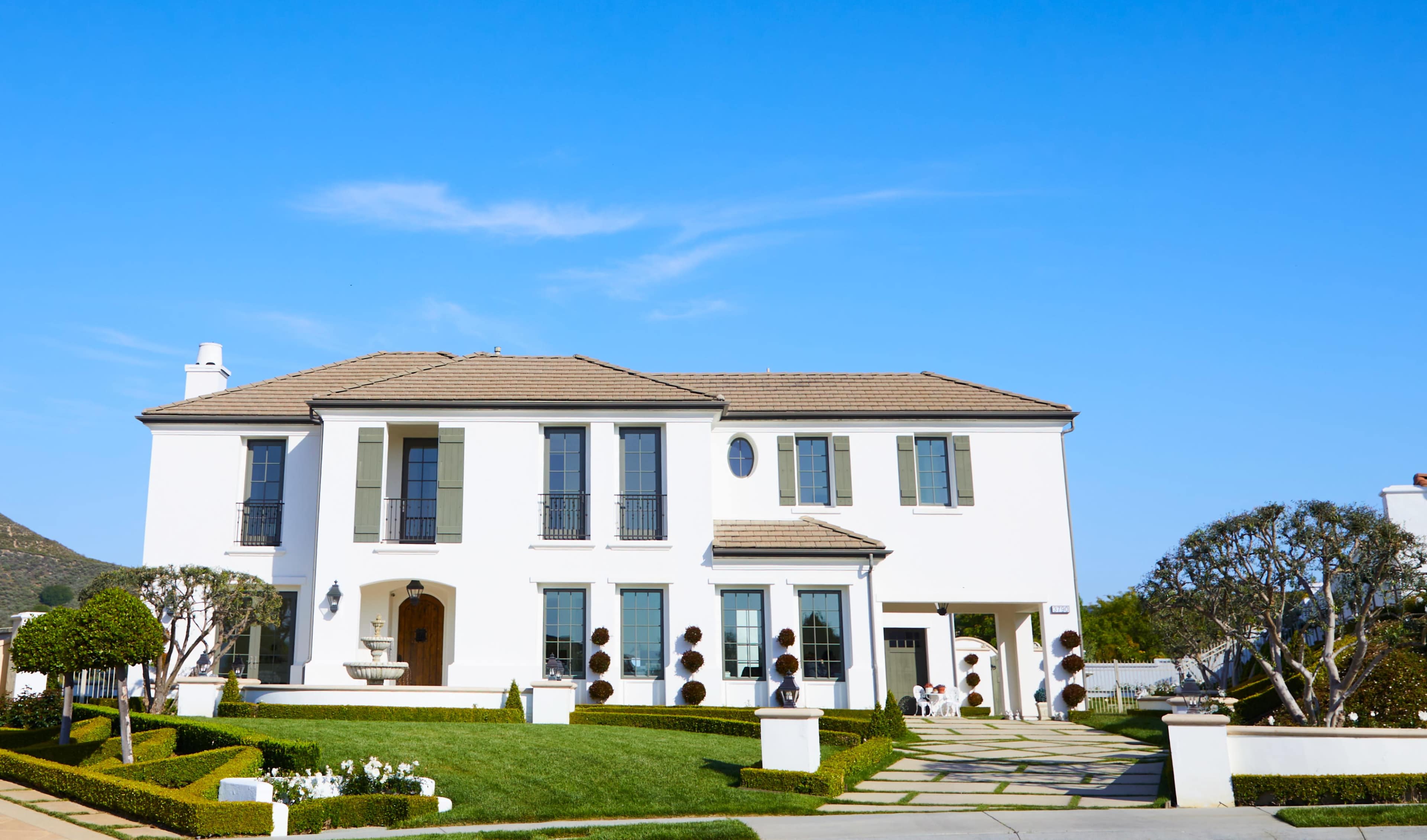 The image shows a two-story white house with green shutters, a tiled roof, and a landscaped front yard featuring bushes and flowers.