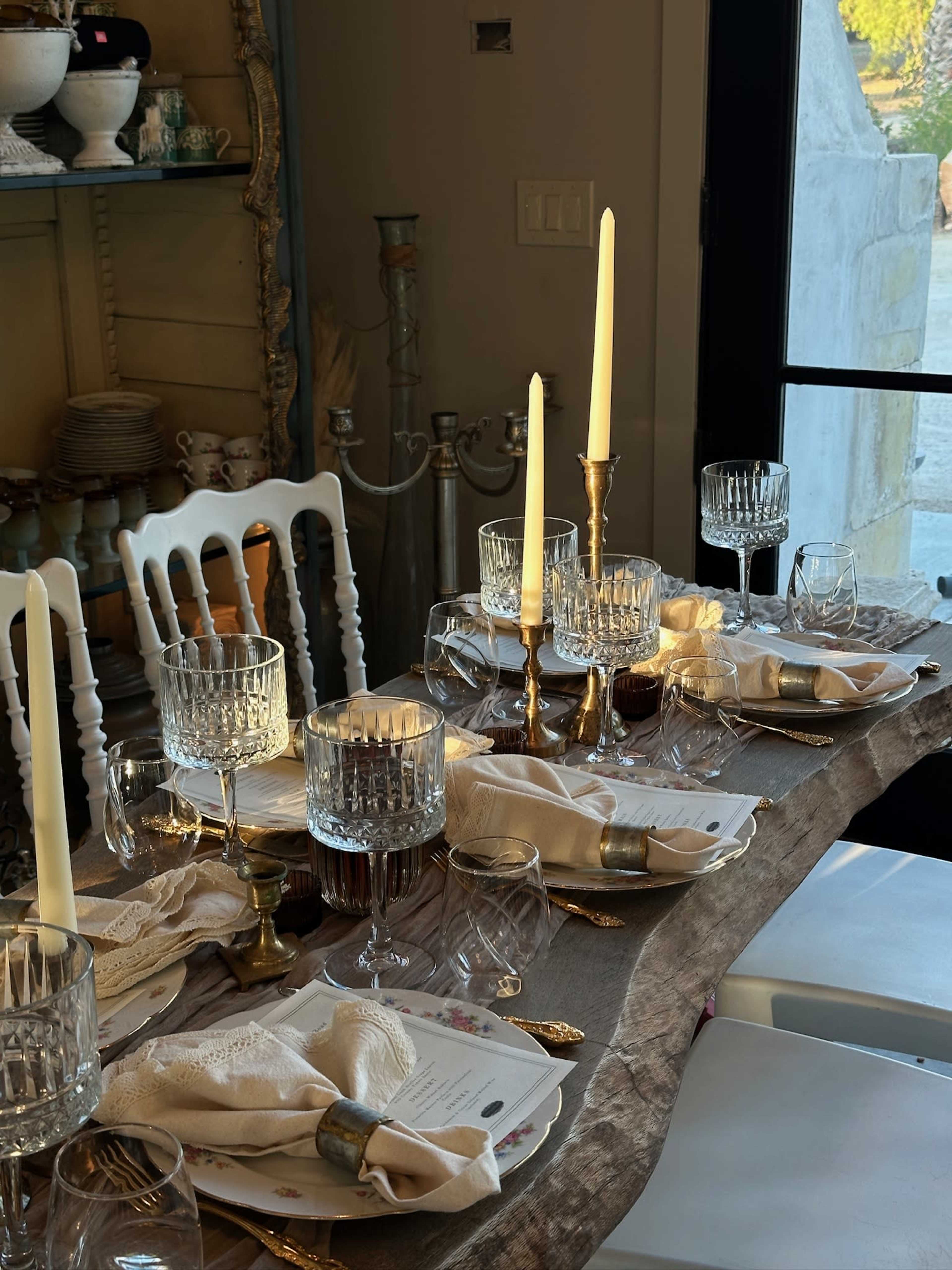 A dining table is set with elegant glassware, silverware, candles, and neatly arranged napkins in a well-lit room.