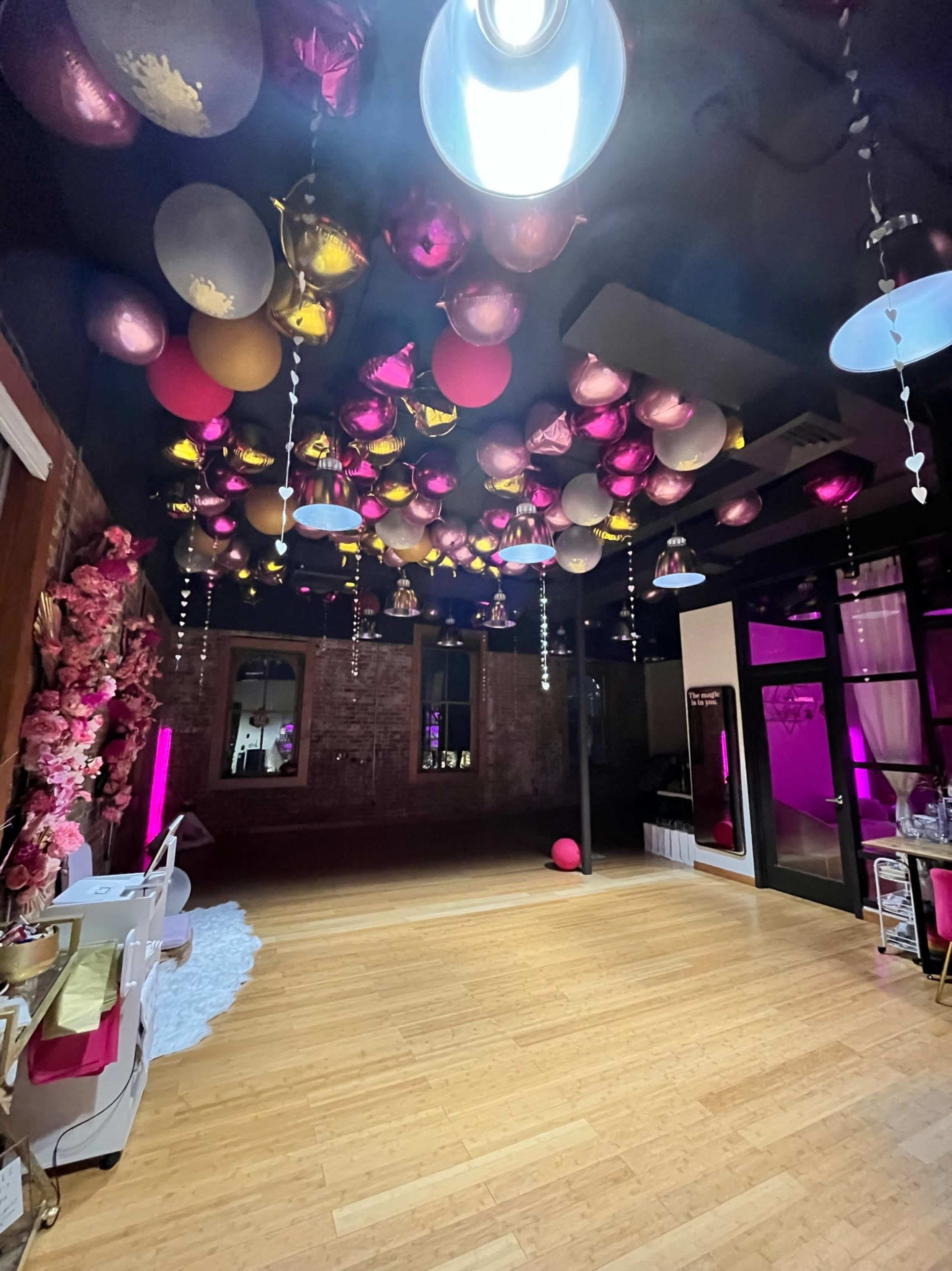 A room with a wooden floor and a ceiling decorated with pink, gold, and silver balloons, along with hanging lights and floral arrangements.