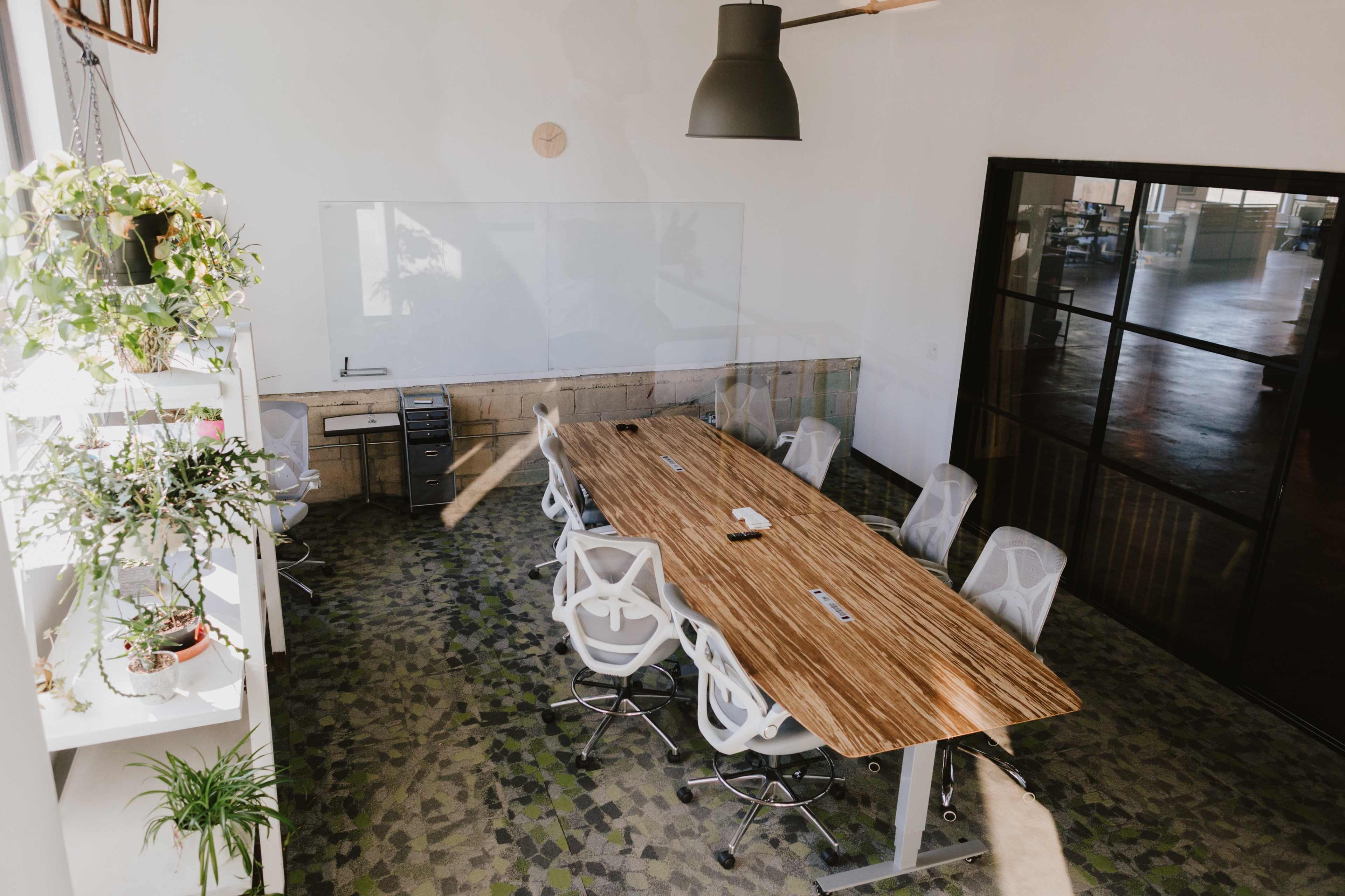 A large conference room features a wooden table surrounded by white office chairs, with a wall-mounted whiteboard and plants in the background.