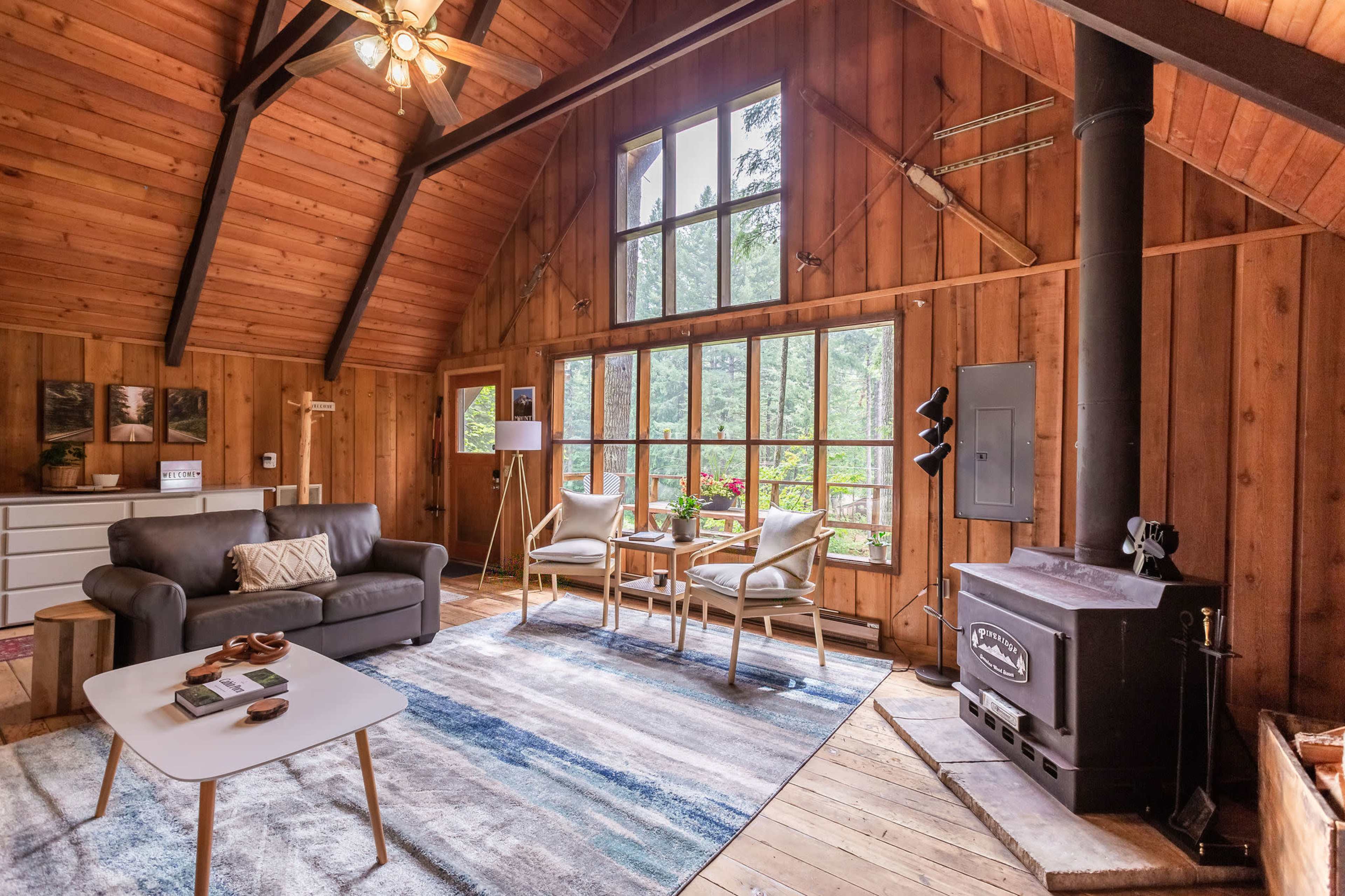 A cozy living room with wooden walls, large windows, a sofa, two chairs, and a wood-burning stove.