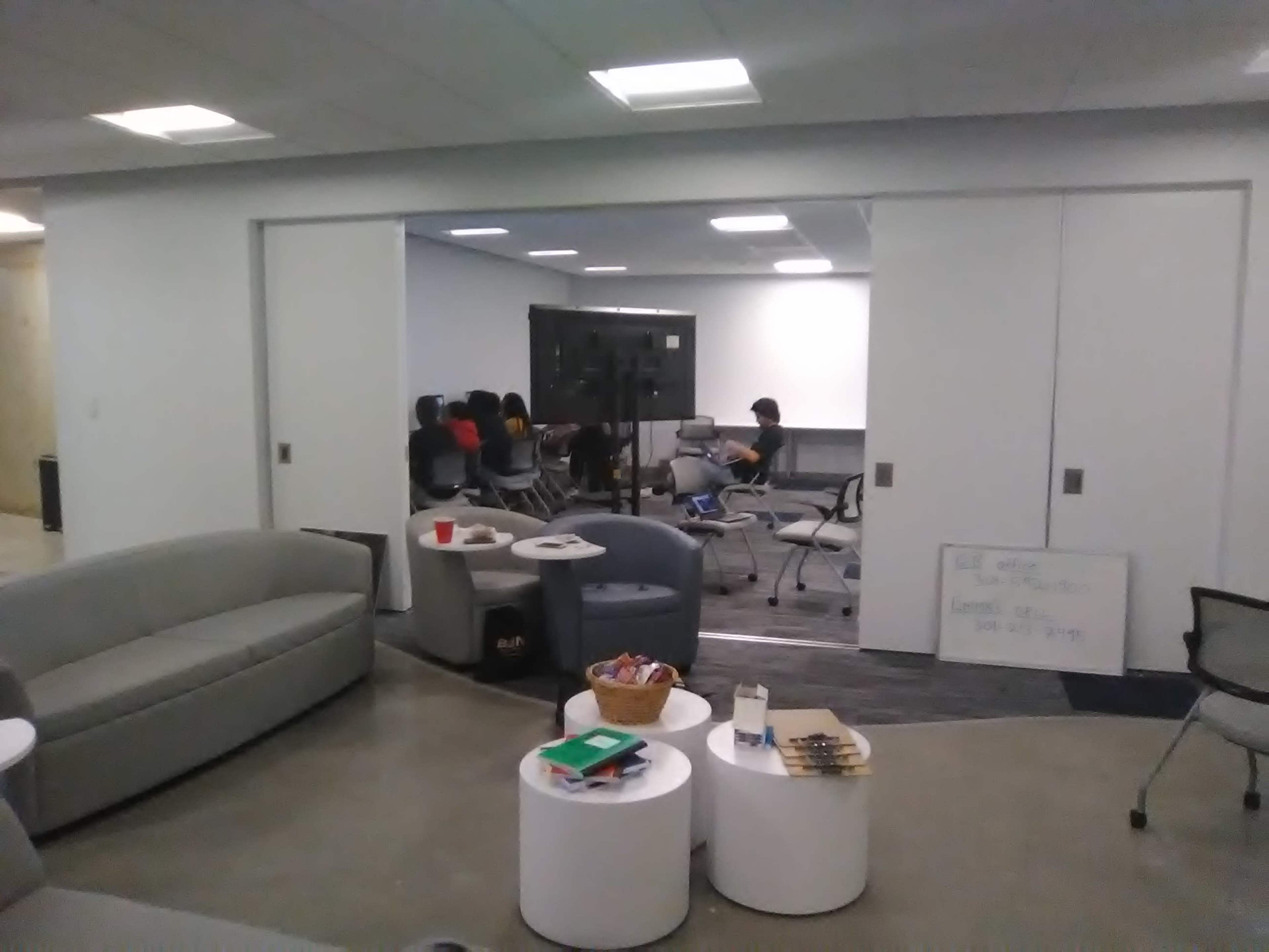 A group of people is seated in a conference room while a seating area with couches and tables is visible in the foreground.