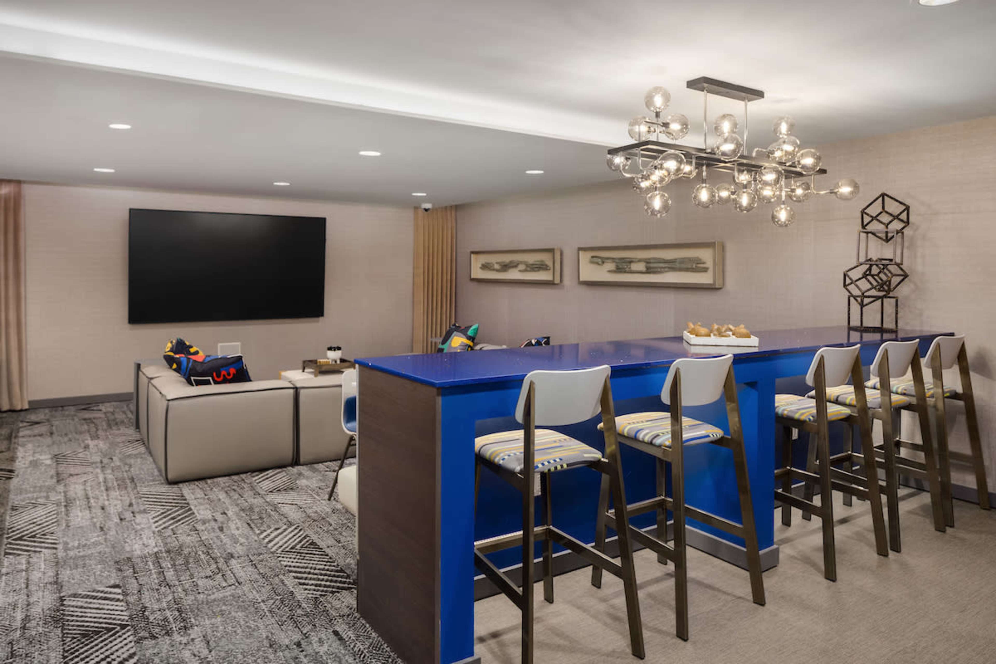 A modern lounge area features a blue countertop bar with high stools, a large wall-mounted TV, and contemporary decor.