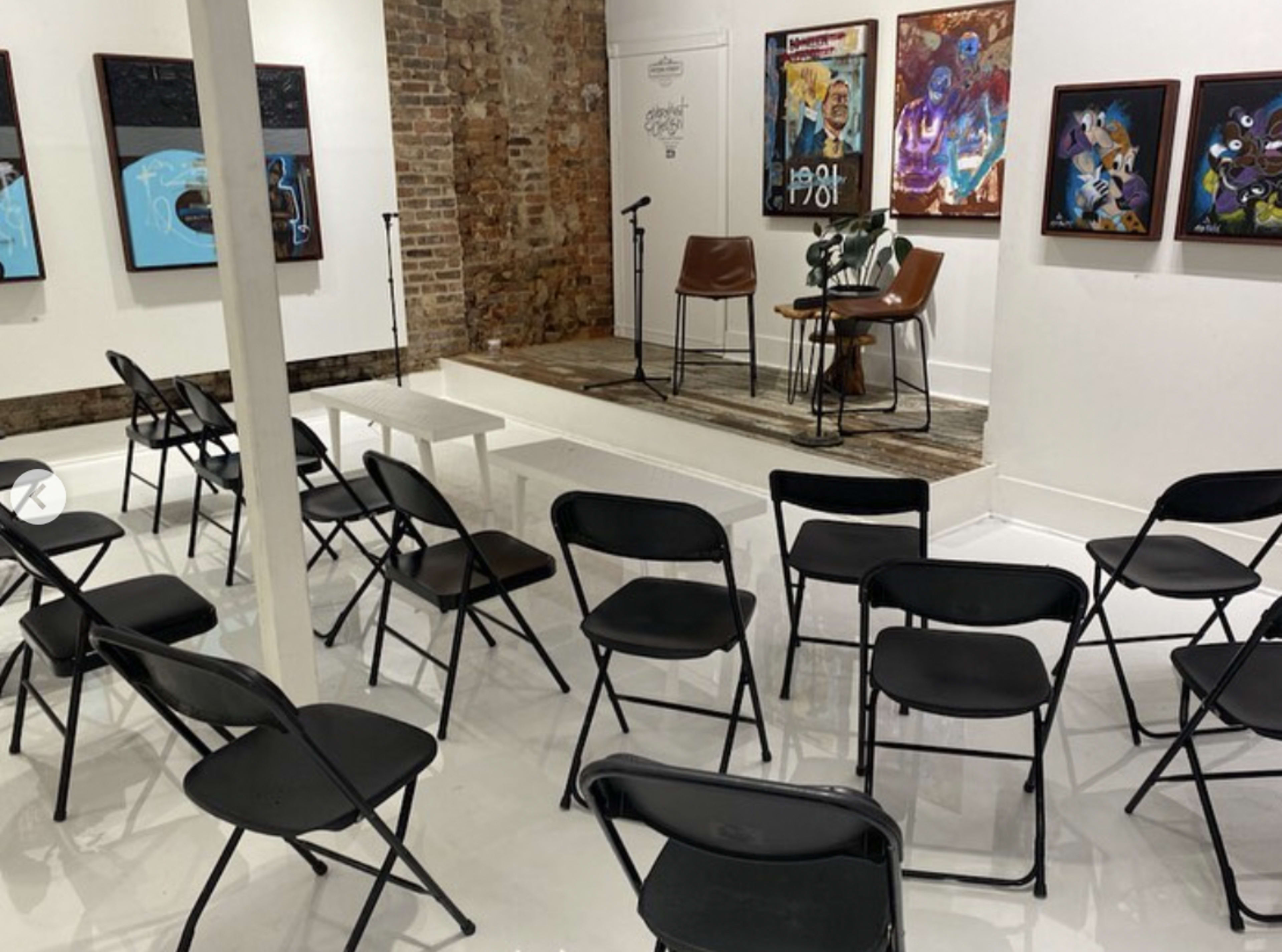 A spacious room with a small stage, two chairs, and several rows of black folding chairs arranged in front of the stage, adorned with artwork on the walls.