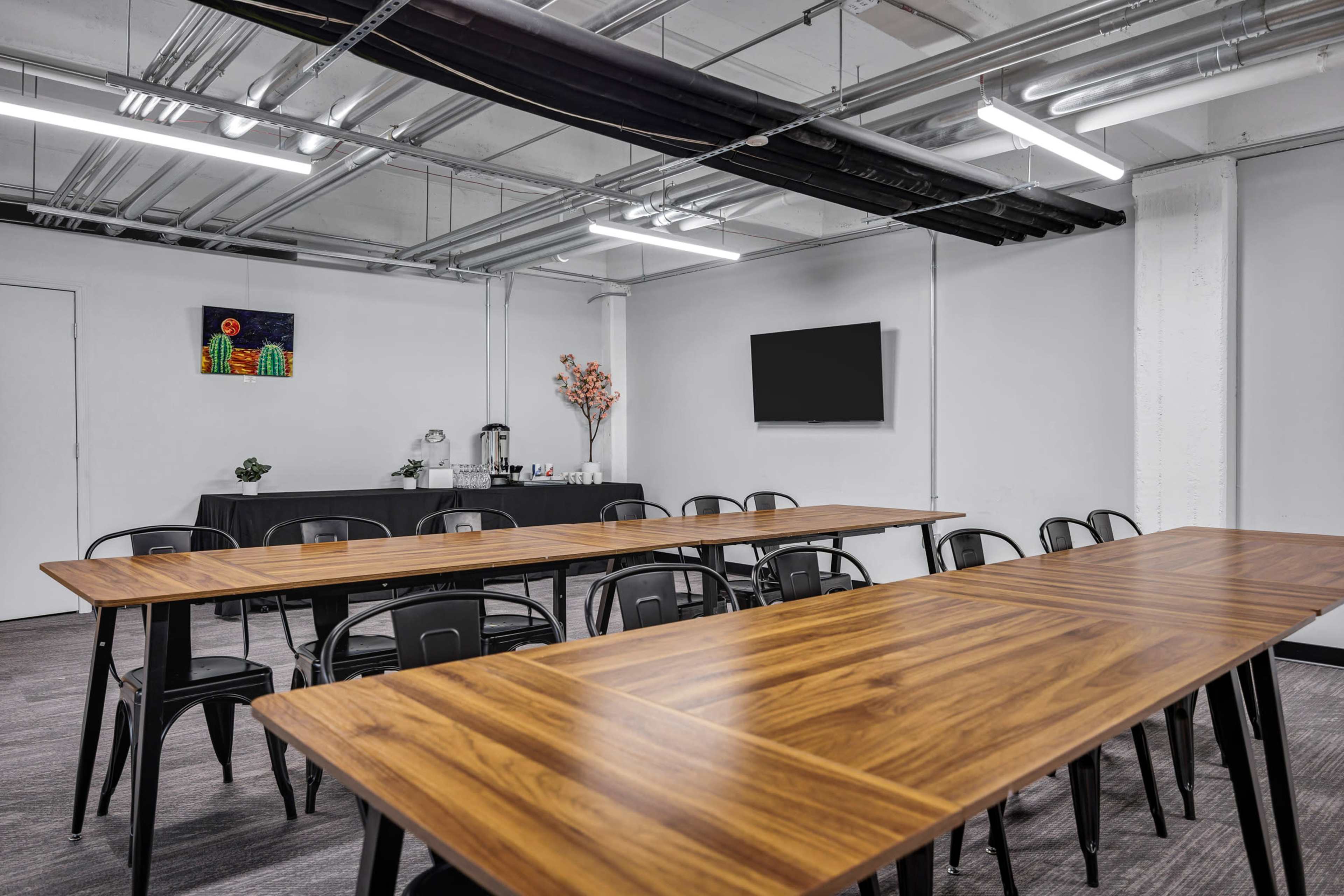 The image shows a modern meeting room with several long wooden tables arranged in rows and black chairs, accompanied by a television on the wall and a refreshment area in the background.