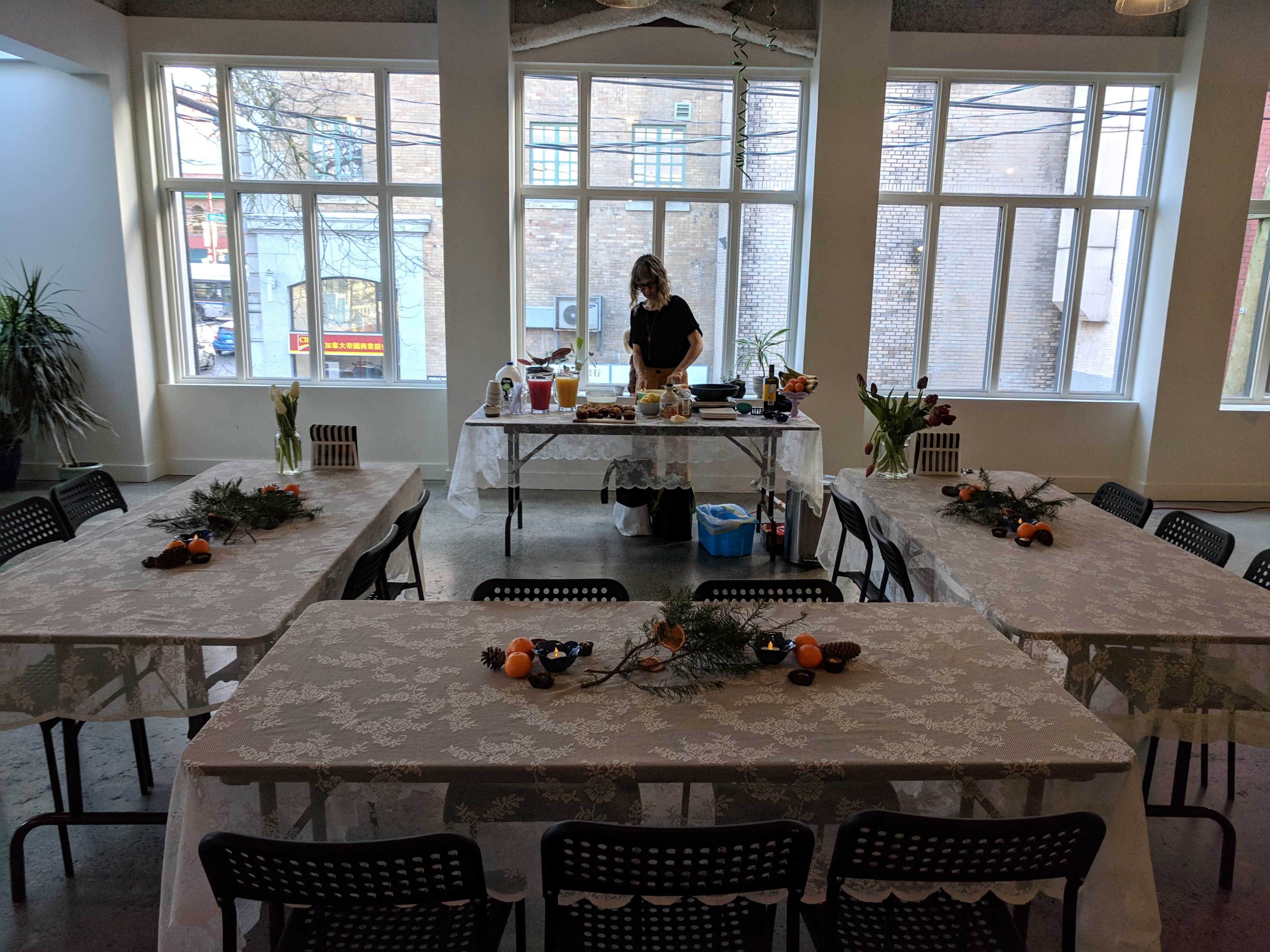 A woman prepares food at a central table surrounded by multiple decorated tables in a brightly lit room with large windows.