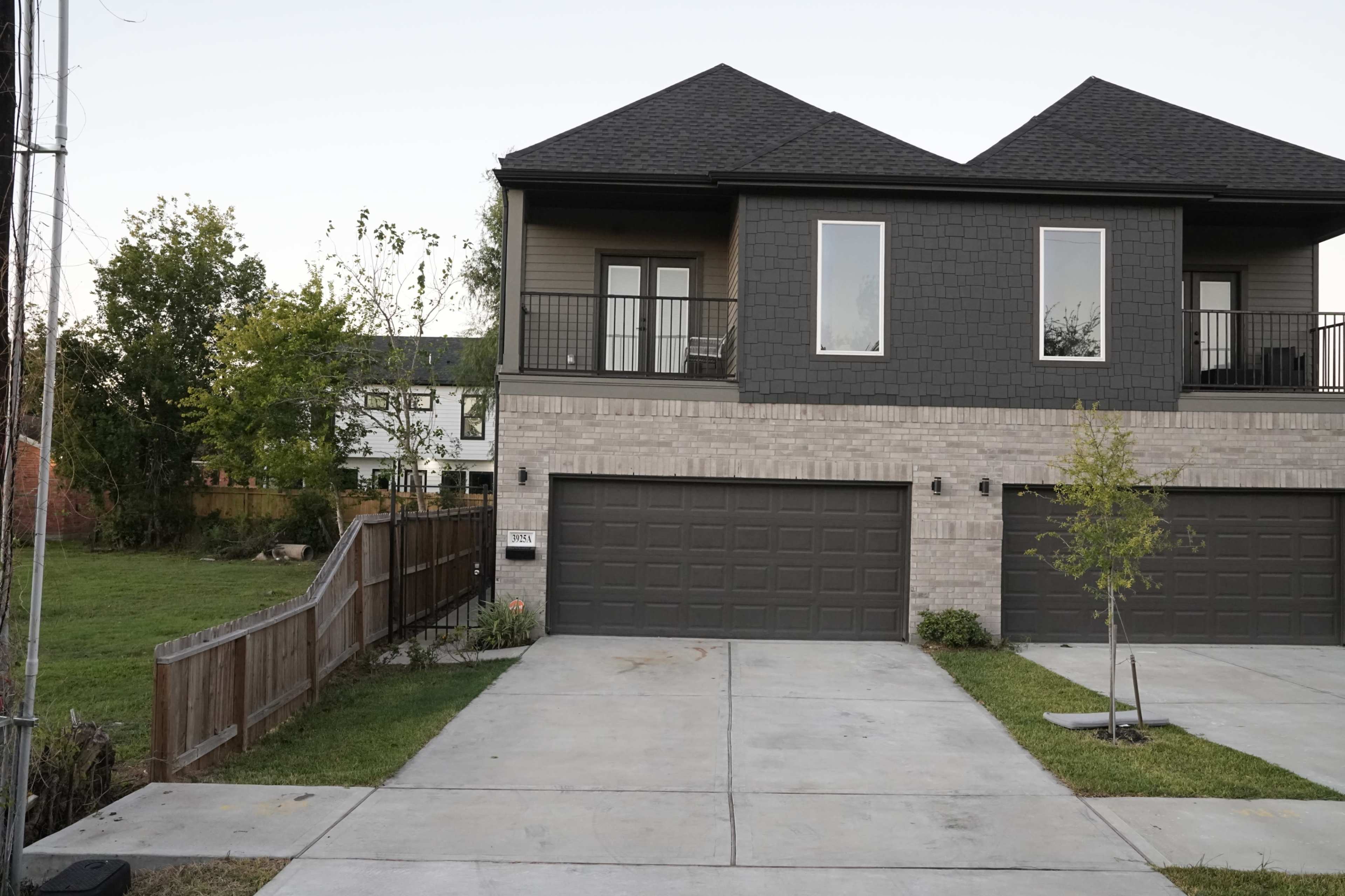 The image shows a two-story duplex with a dark-colored upper section and brick lower section, featuring a driveway and a fenced yard.