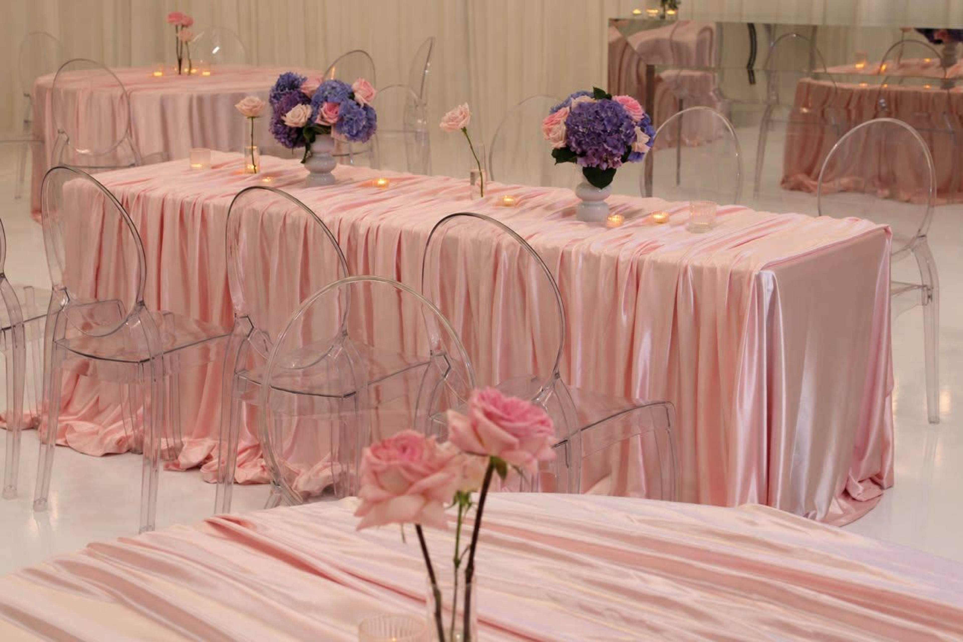 The image shows tables covered with pink satin cloths and adorned with floral arrangements and candles in a softly lit venue.