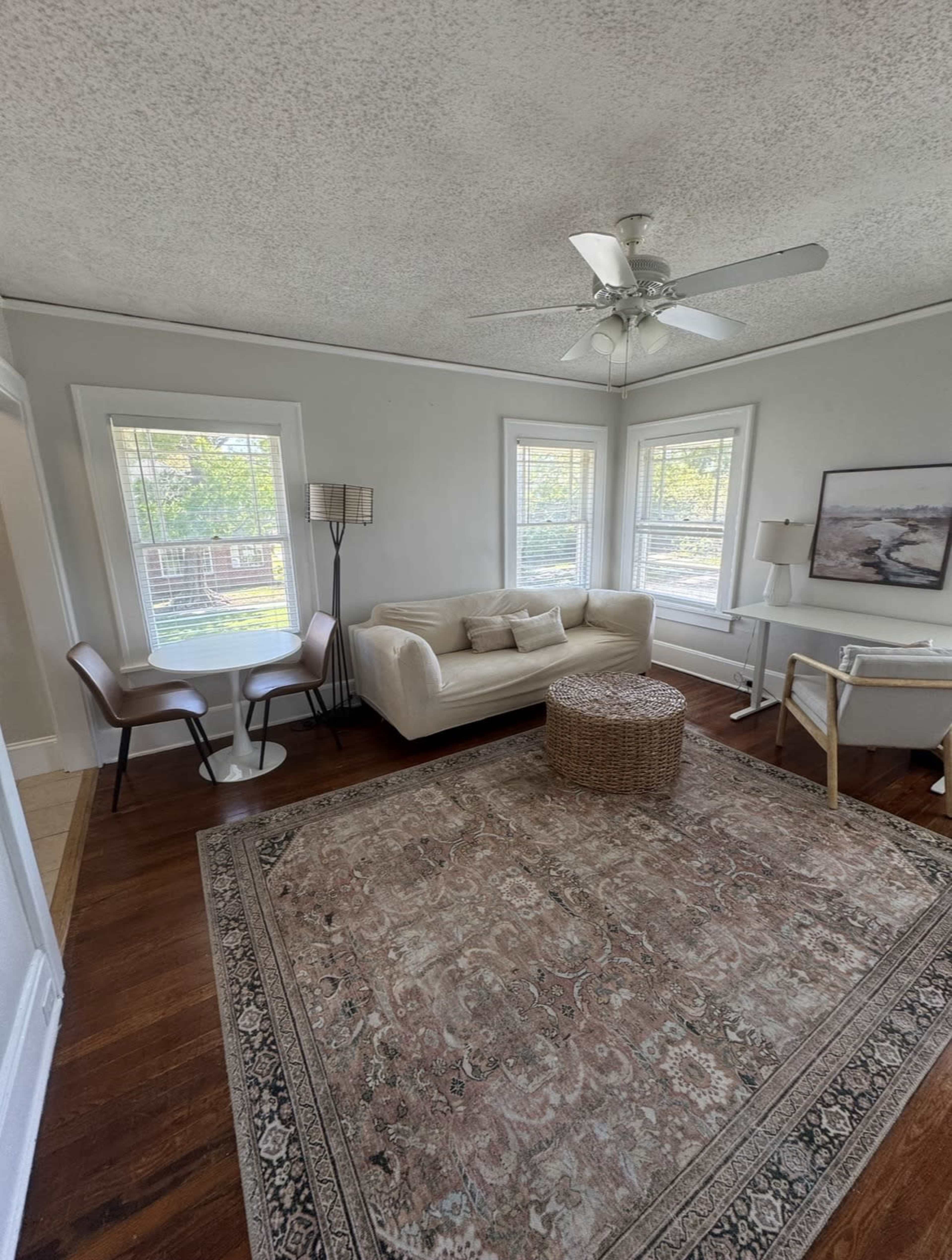 The image shows a living room with a beige sofa, a round woven coffee table, and two chairs next to a window, all set on a patterned rug.