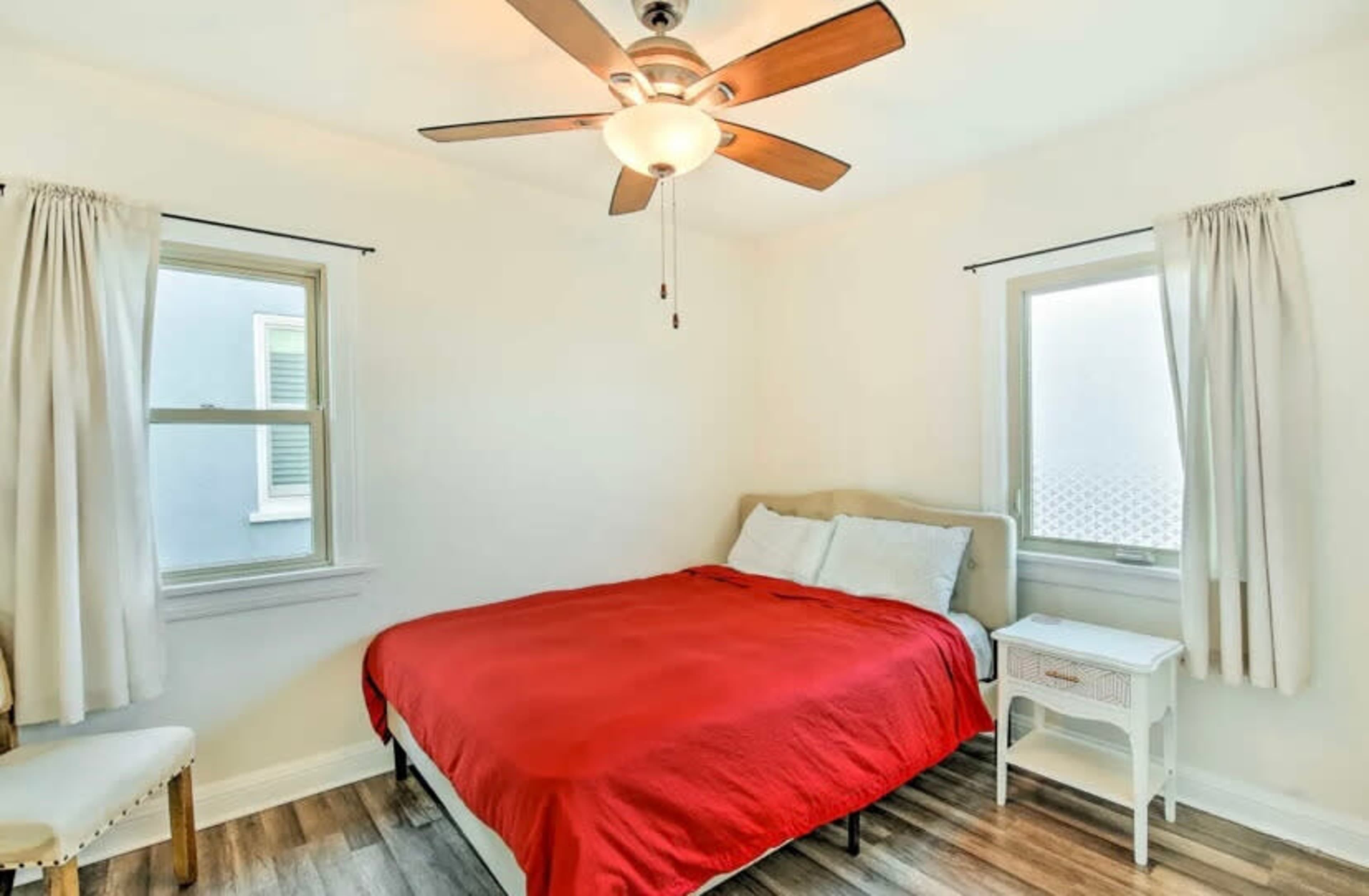 A brightly lit bedroom featuring a bed with a red bedspread, a ceiling fan, and two windows.