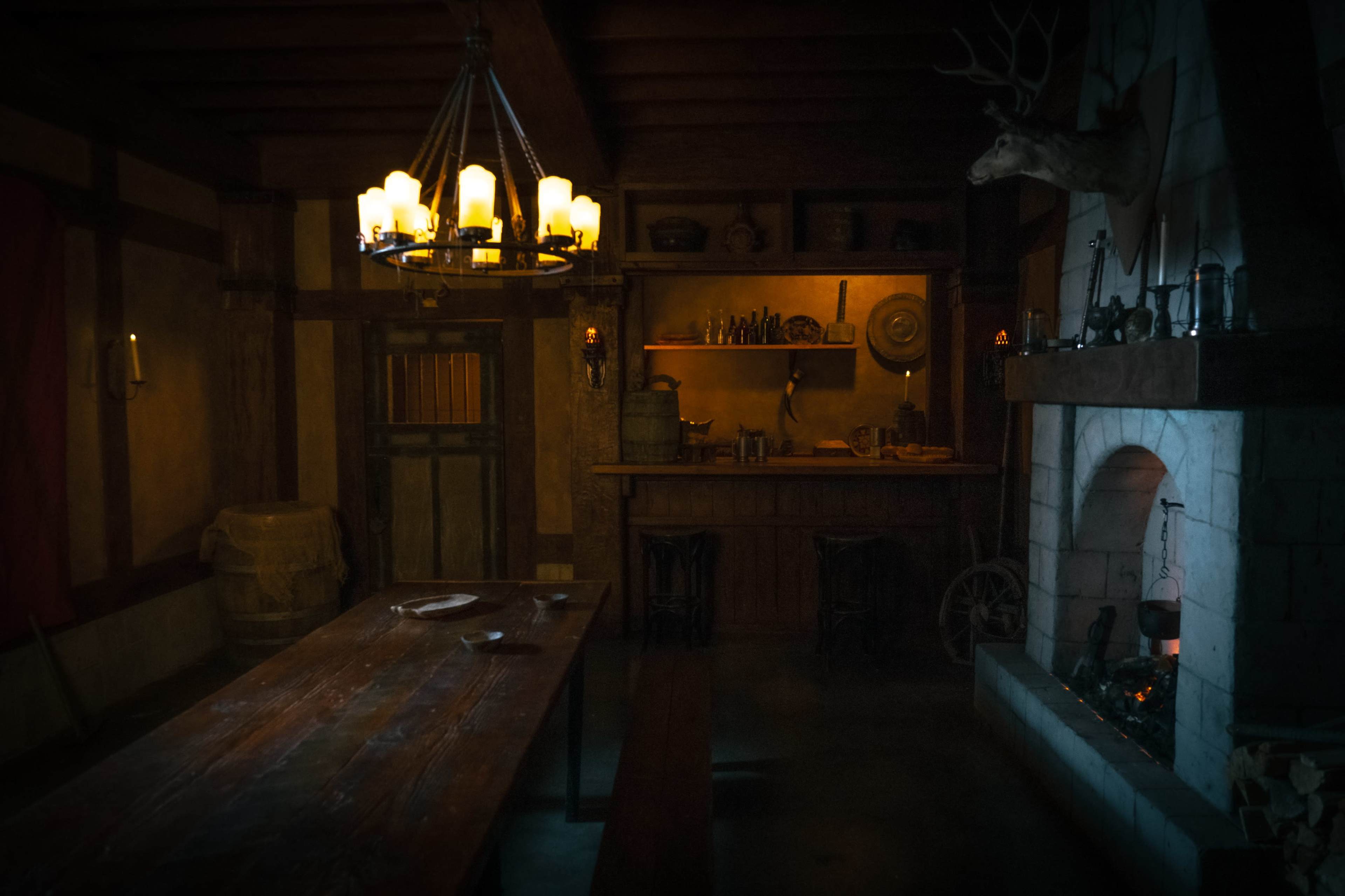 The image shows a dimly lit rustic interior of a tavern or dining room with a large wooden table, a central chandelier, and a stone fireplace.