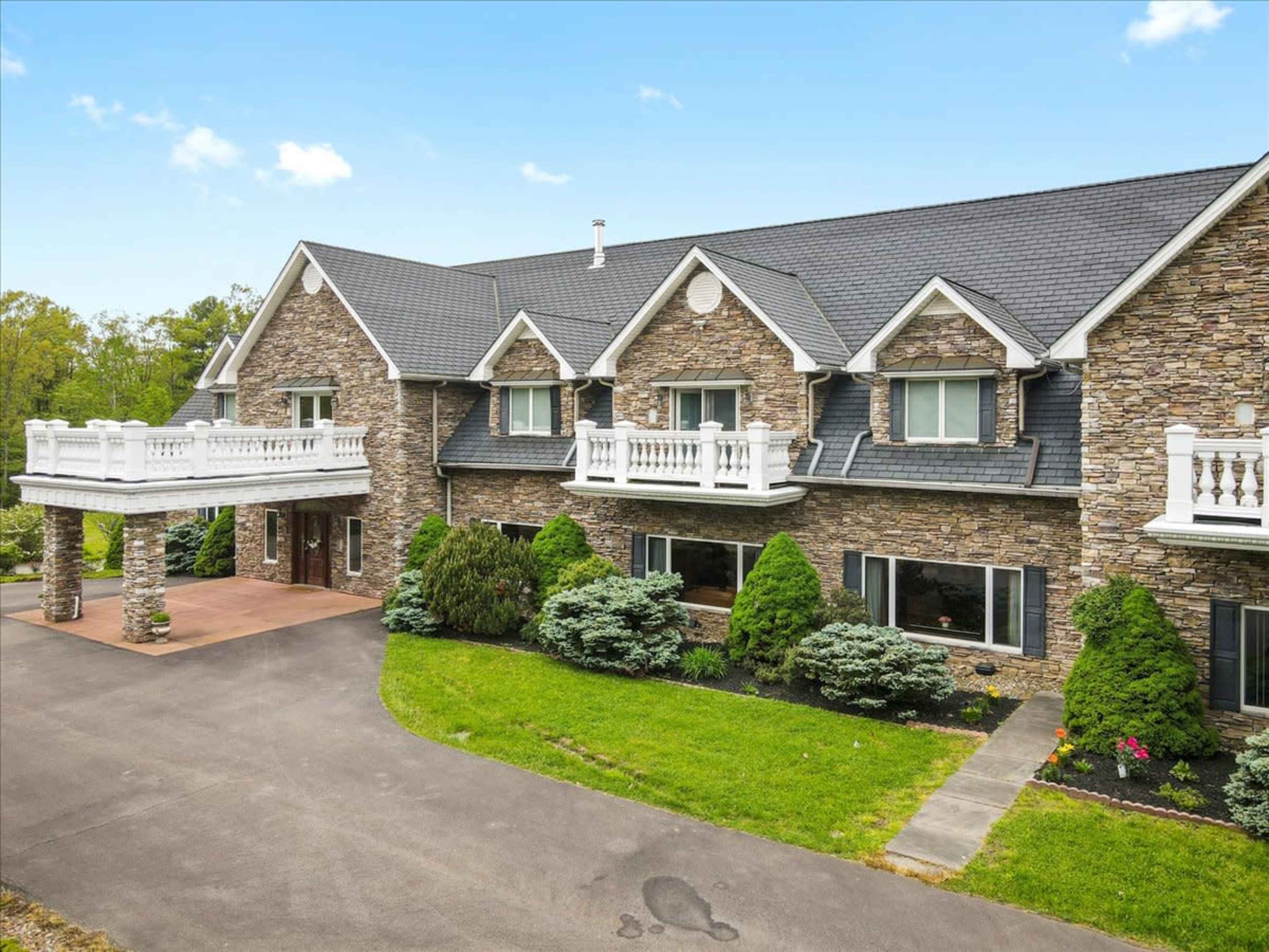 A large, stone-faced house with multiple gabled roofs, surrounded by manicured greenery and a paved driveway.