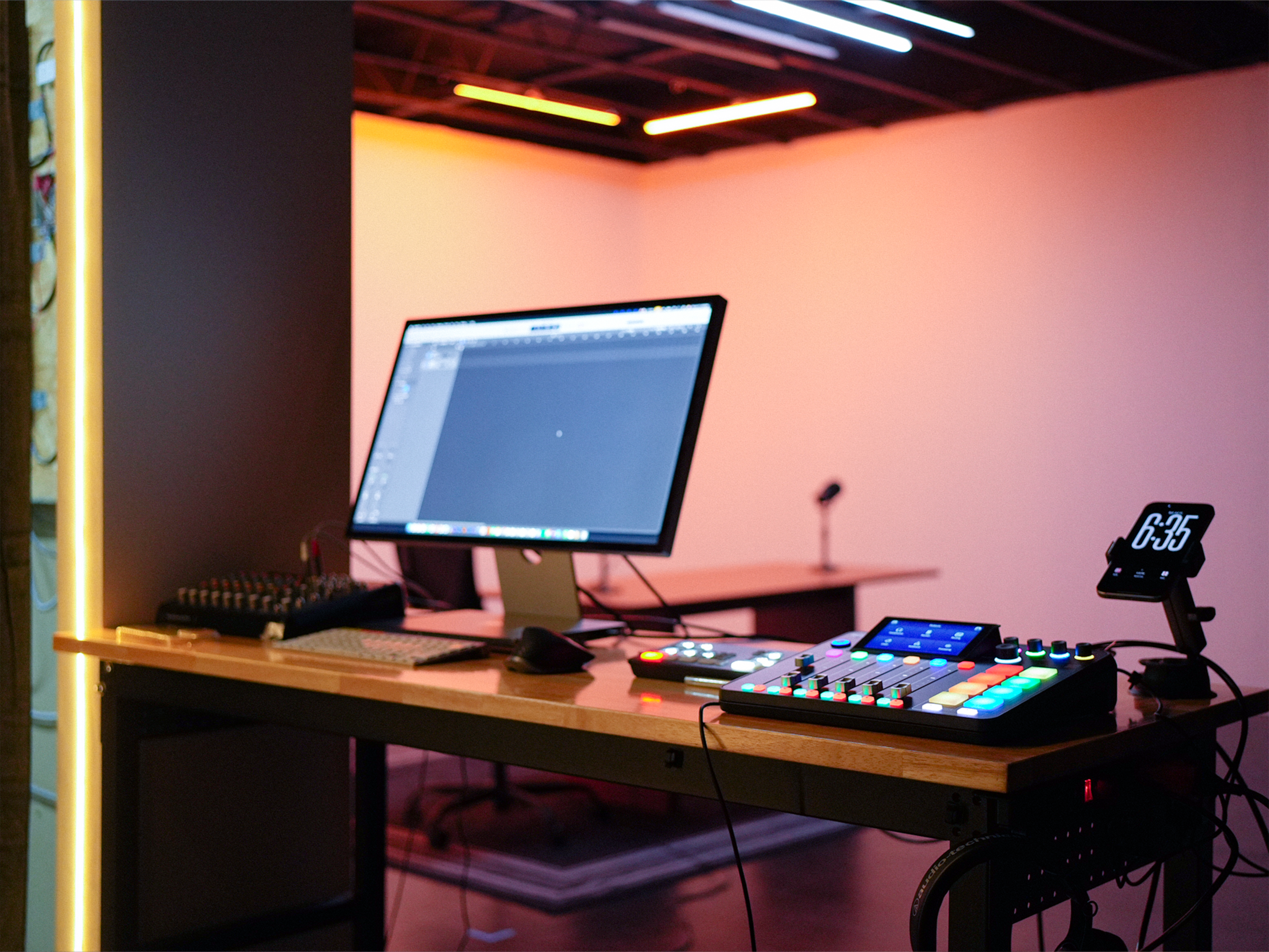 A computer monitor is positioned next to a soundboard and control interface on a table in a dimly lit room with neon lights.