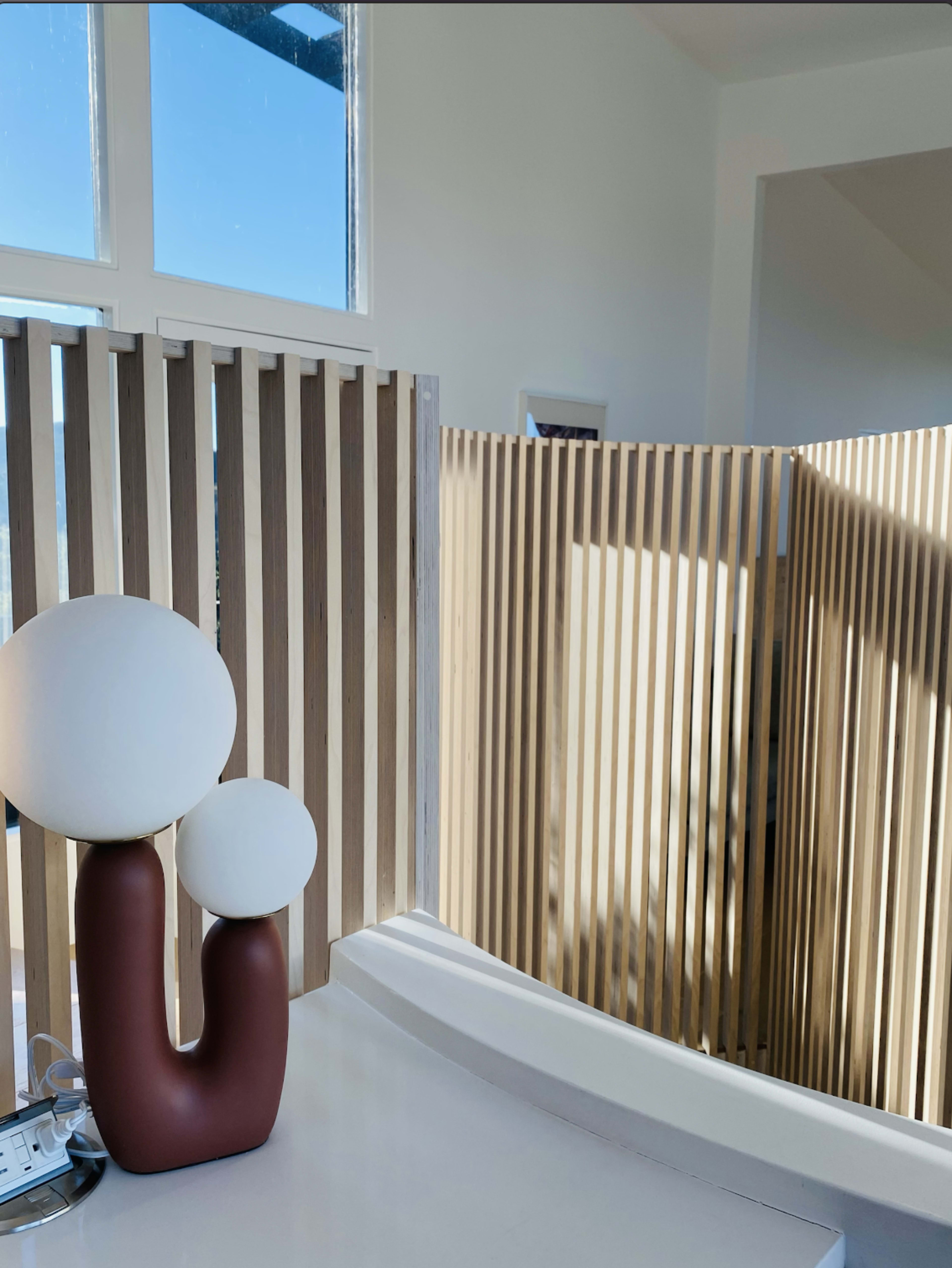 A modern interior scene with a brown and white table lamp beside a wooden slatted partition and large windows allowing natural light to enter.