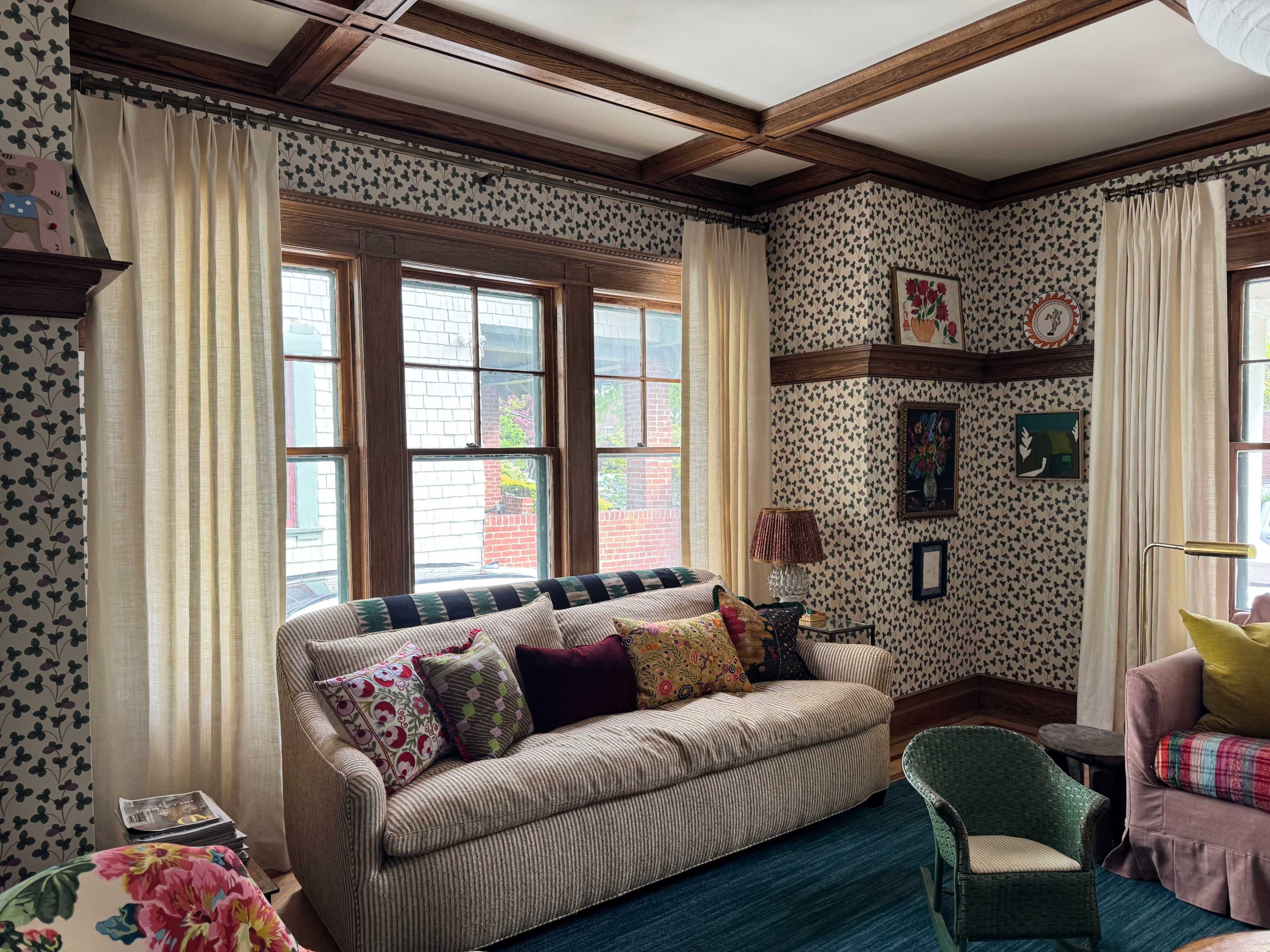 The image shows a cozy living room with patterned wallpaper, large windows, and a sofa adorned with colorful cushions.