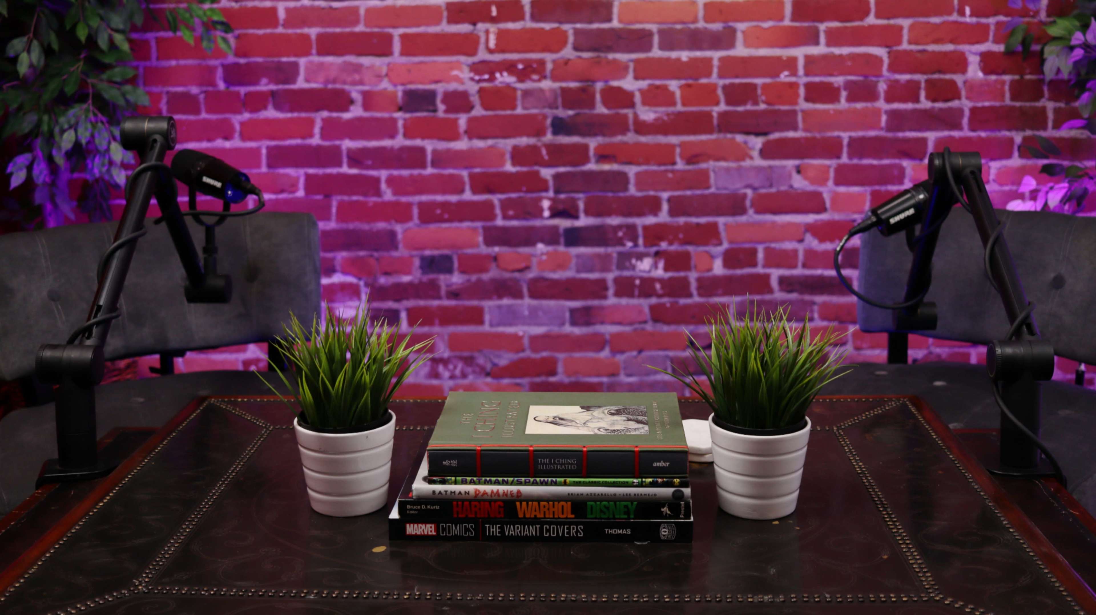 The image shows a small table with stacked books and potted plants between two microphone stands against a brick wall with purple lighting.