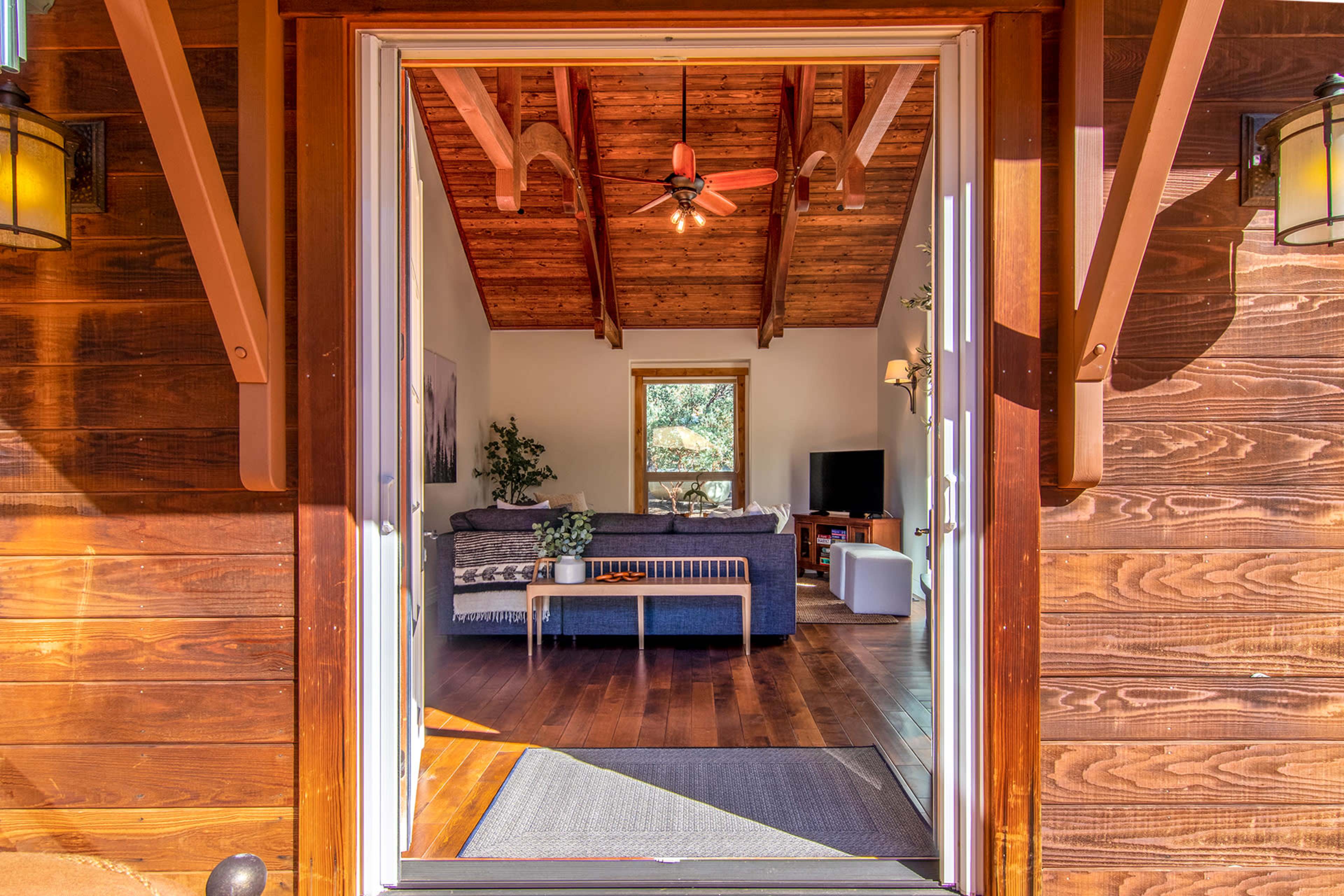 A view from the entrance of a wooden cabin showing a living area with a couch, coffee table, and large windows.