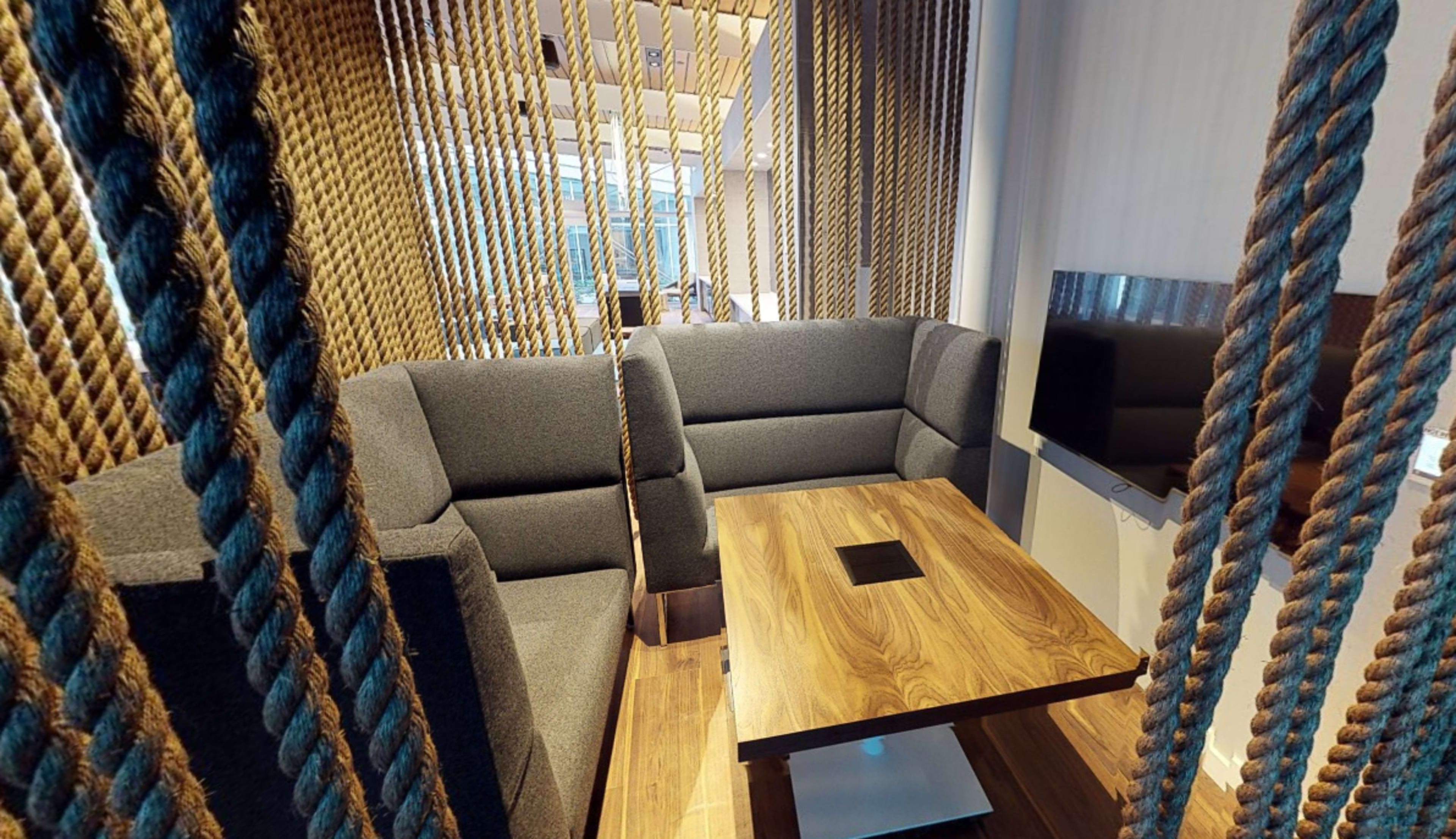 The image shows a modern seating area enclosed by thick ropes, featuring gray upholstered sofas and a wooden table with a central power outlet.