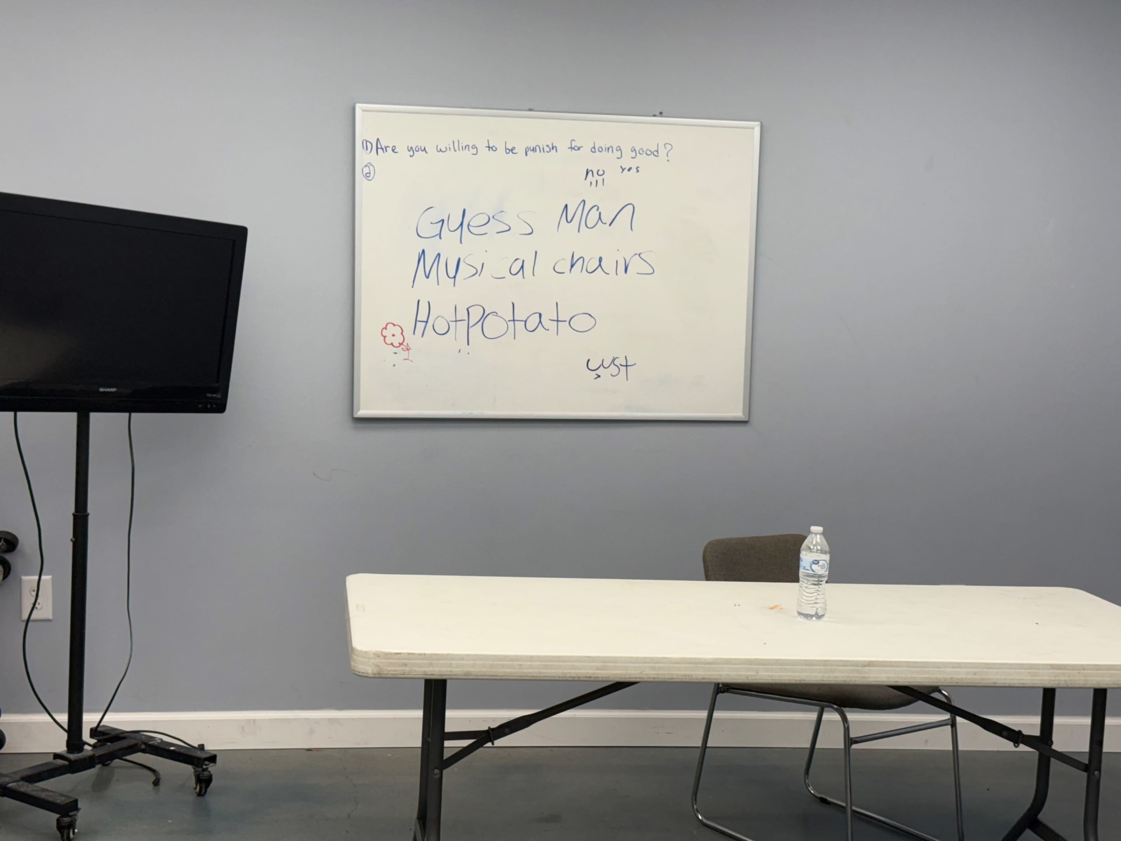 A whiteboard with handwritten activity titles, including "Guess Man," "Musical Chairs," and "Hot Potato," is displayed beside a table and a chair in a simple meeting room.