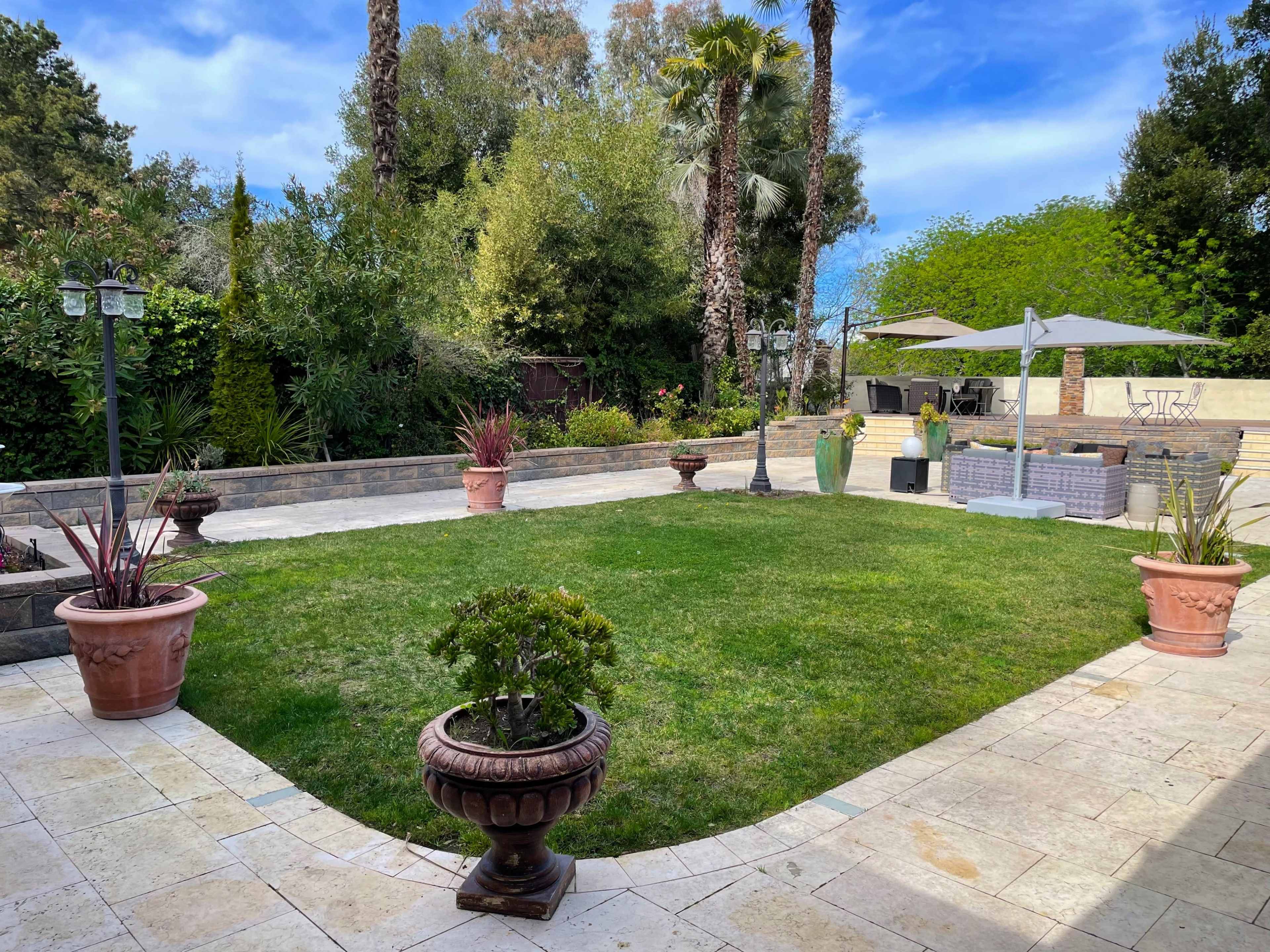 A landscaped garden with a grassy area surrounded by stone paths, decorative plants in pots, and outdoor furniture under a patio umbrella.