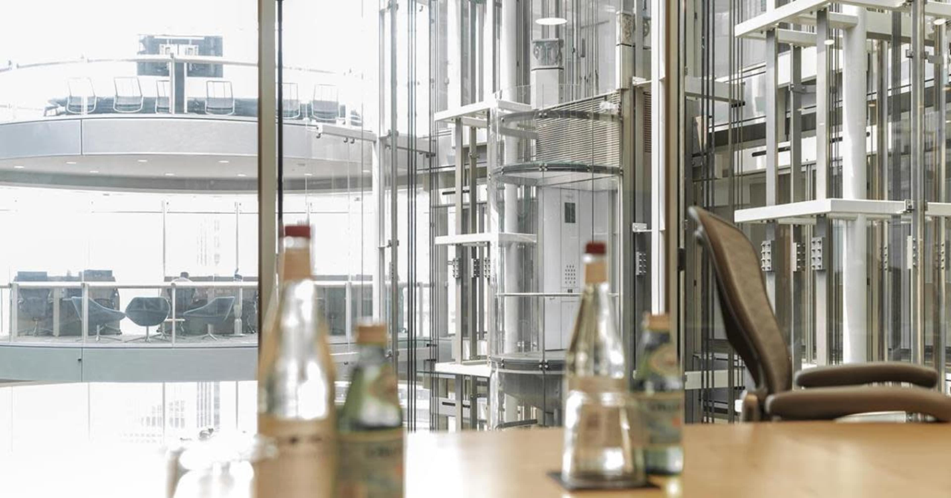 A conference room with a table featuring several glass bottles and a view of an elevator shaft and meeting area through large glass windows.