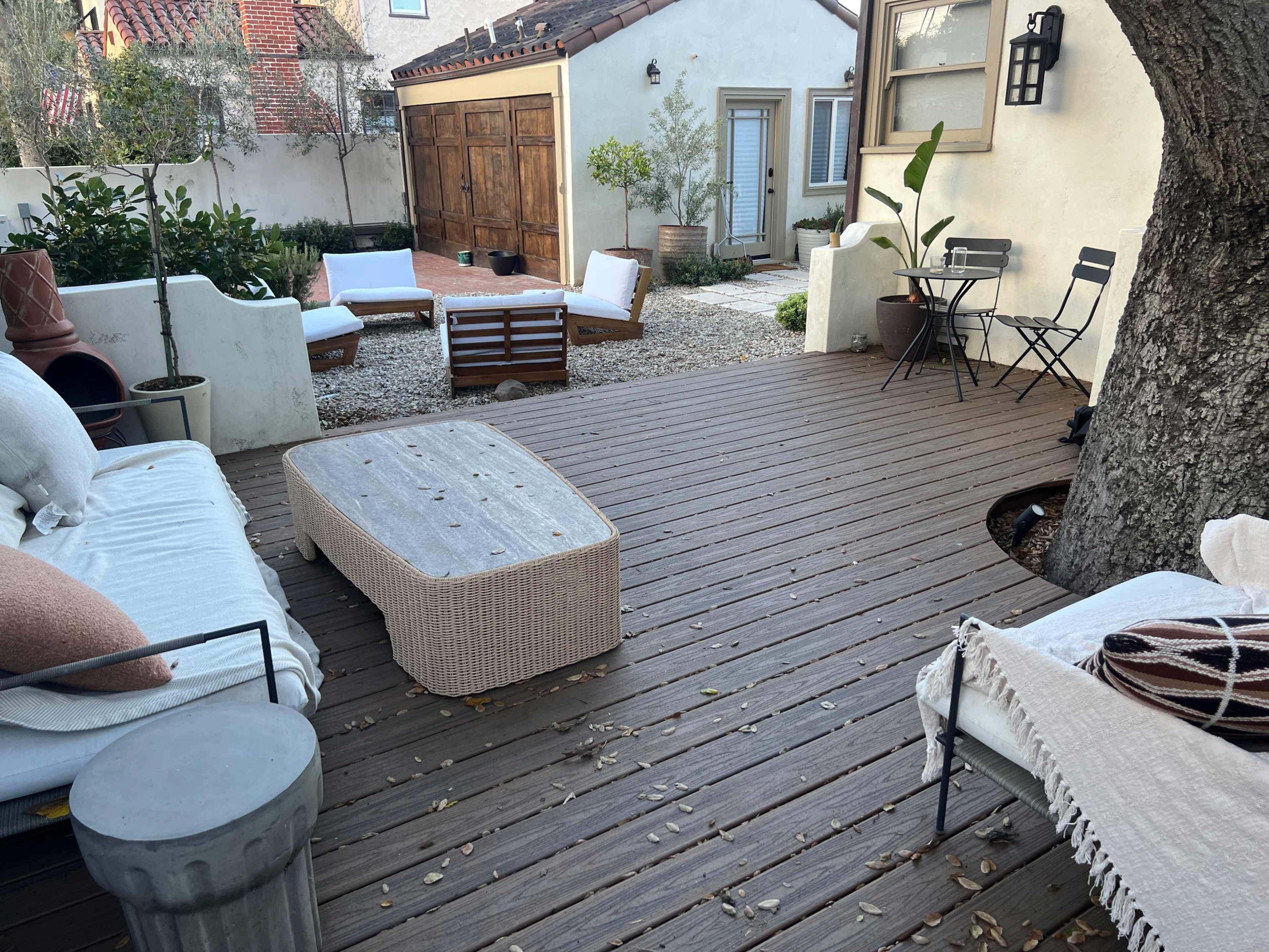 The image shows a patio area with wooden decking, featuring a coffee table, two lounge chairs, and a small bistro set, surrounded by potted plants and a gravel garden.