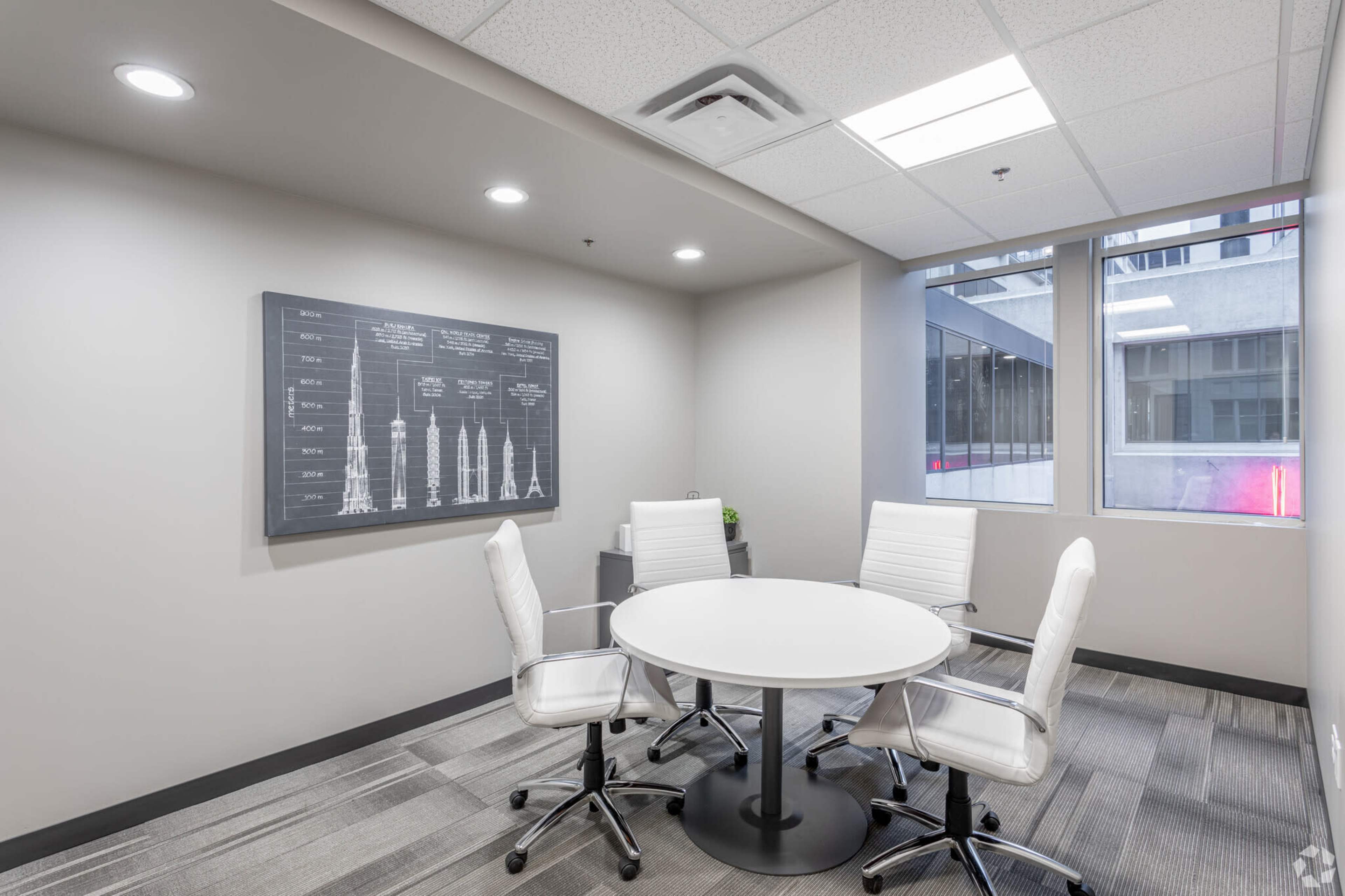 A small meeting room with a round table and four white chairs, accompanied by a black wall chart and large windows.