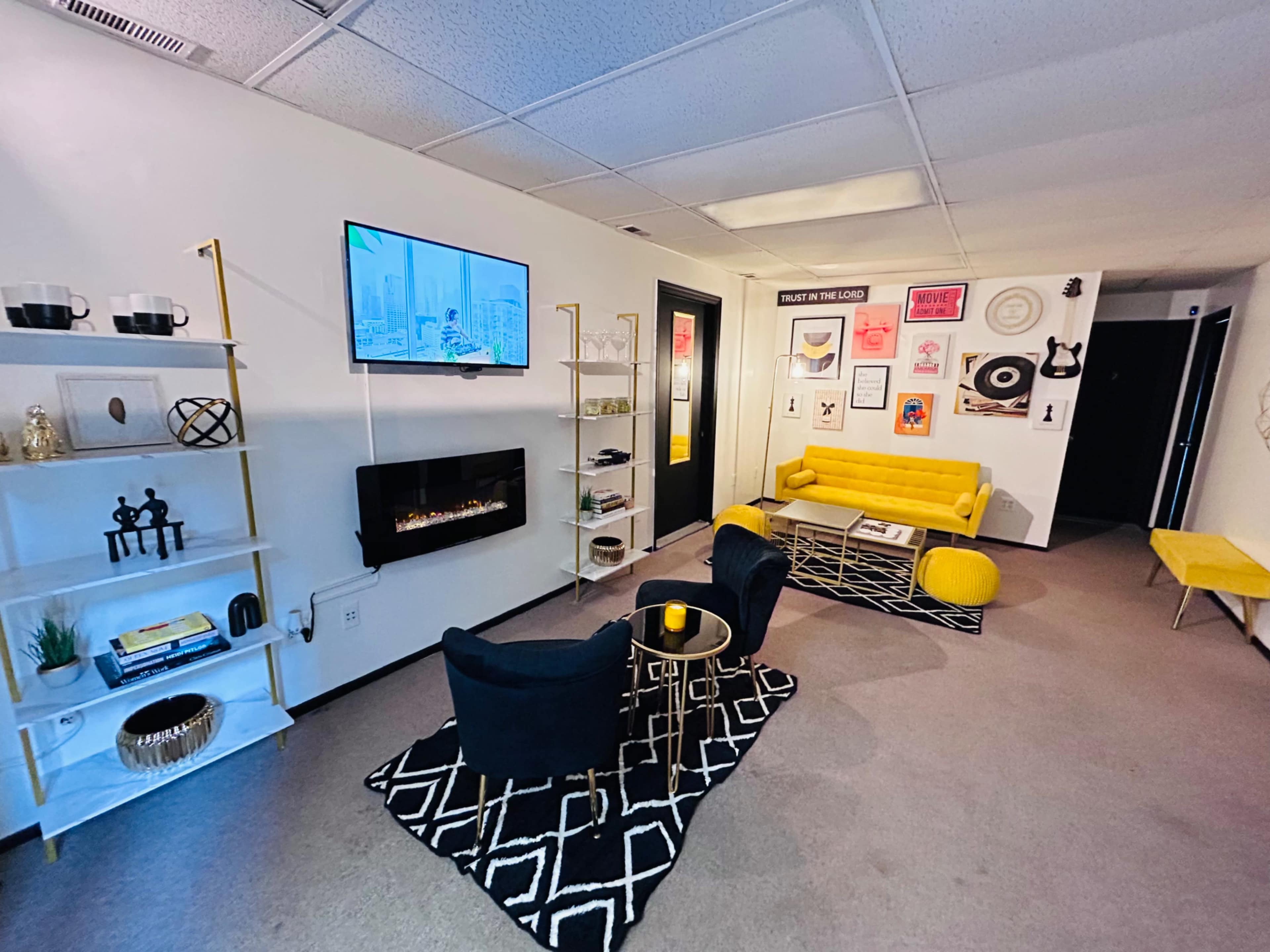 The image shows a modern office space featuring a yellow sofa, black chairs, a wall-mounted TV, and decorative artwork.