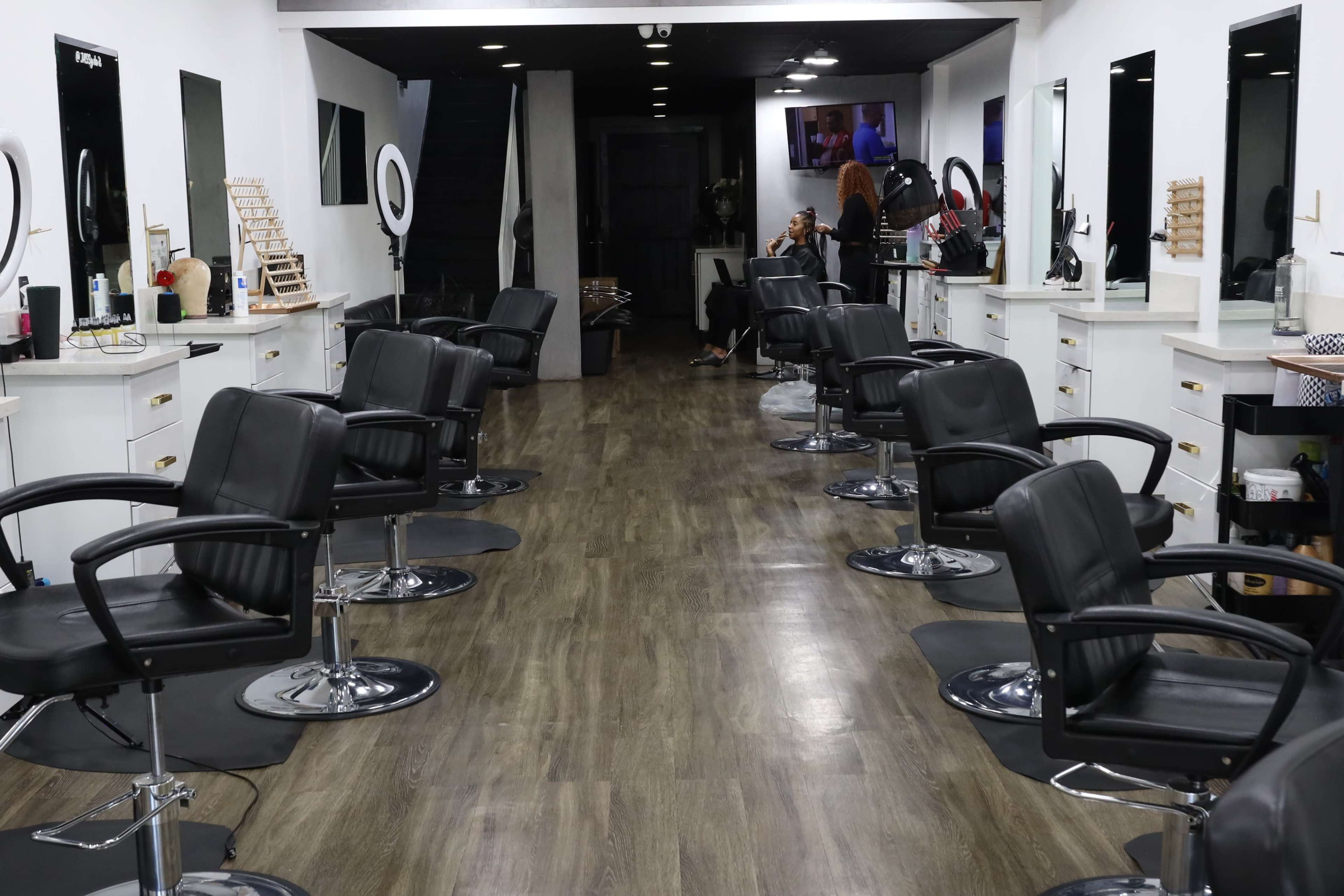 The interior of a salon with empty black styling chairs arranged in a row along a wooden floor, and mirrors mounted on white walls.