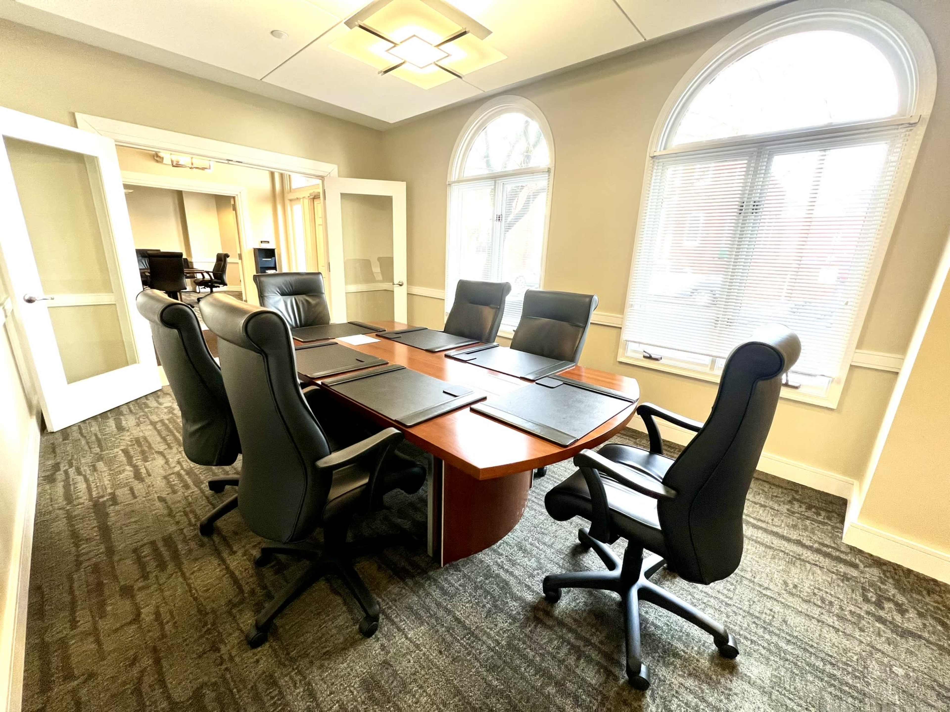 A conference room features a large, oval table surrounded by six black swivel chairs, with large windows illuminating the space.