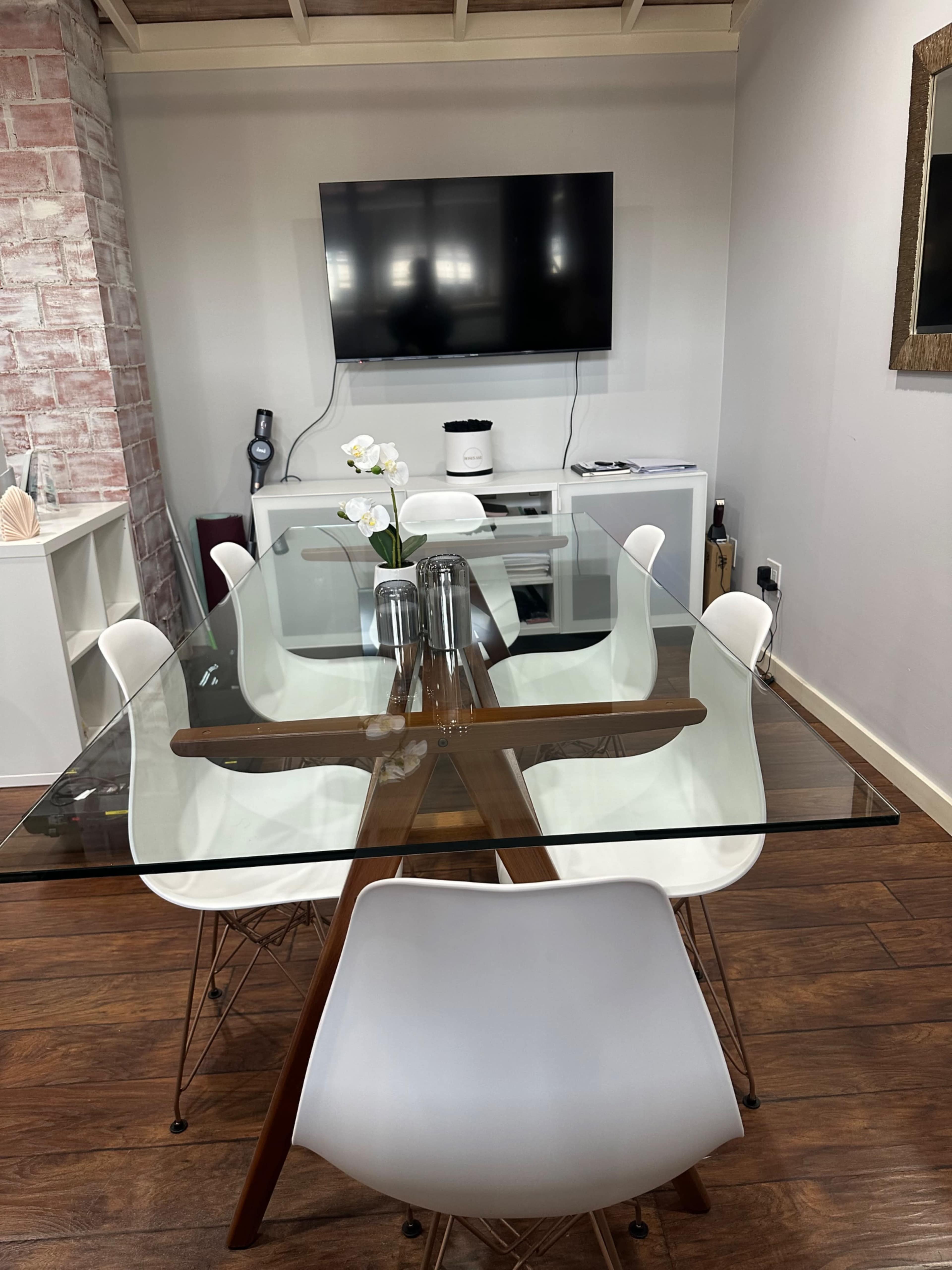 A modern dining room features a glass-top table surrounded by white chairs, with a television mounted on the wall and a cabinet in the background.