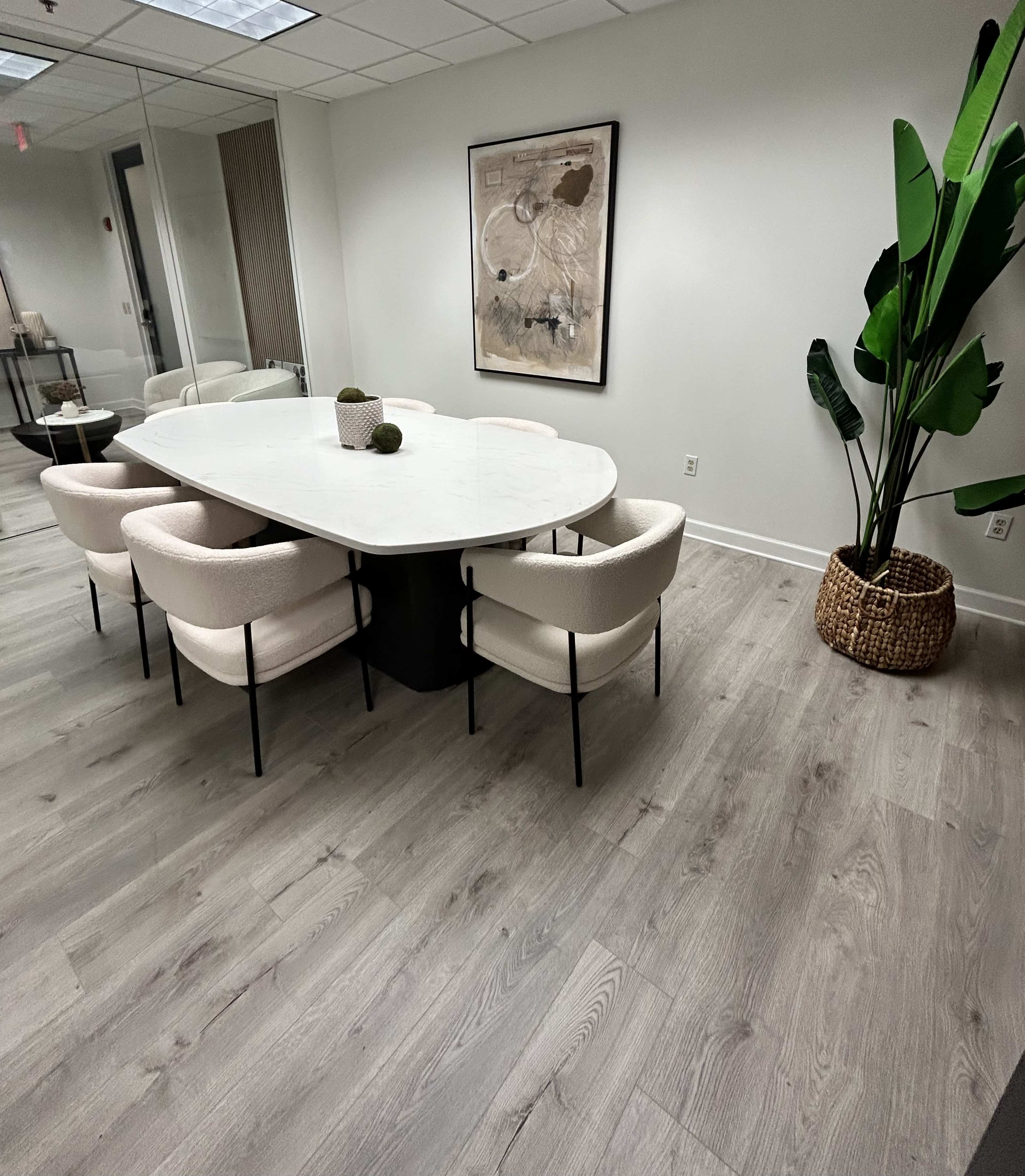 A conference room features a large marble table surrounded by four upholstered chairs, with a potted plant in a woven basket against a wall.