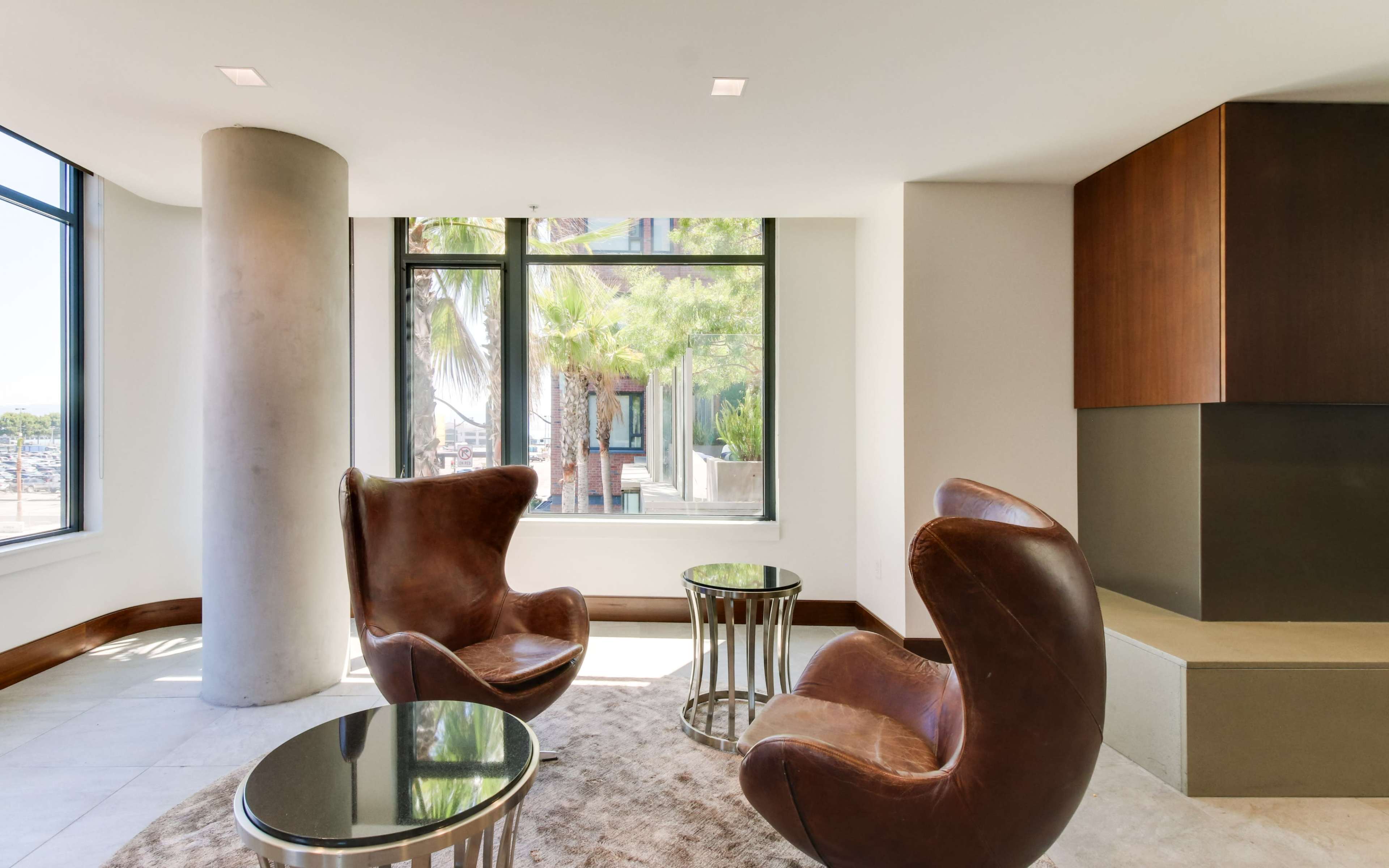 The image shows a modern seating area featuring two brown chairs, small glass tables, and large windows with a view of palm trees outside.