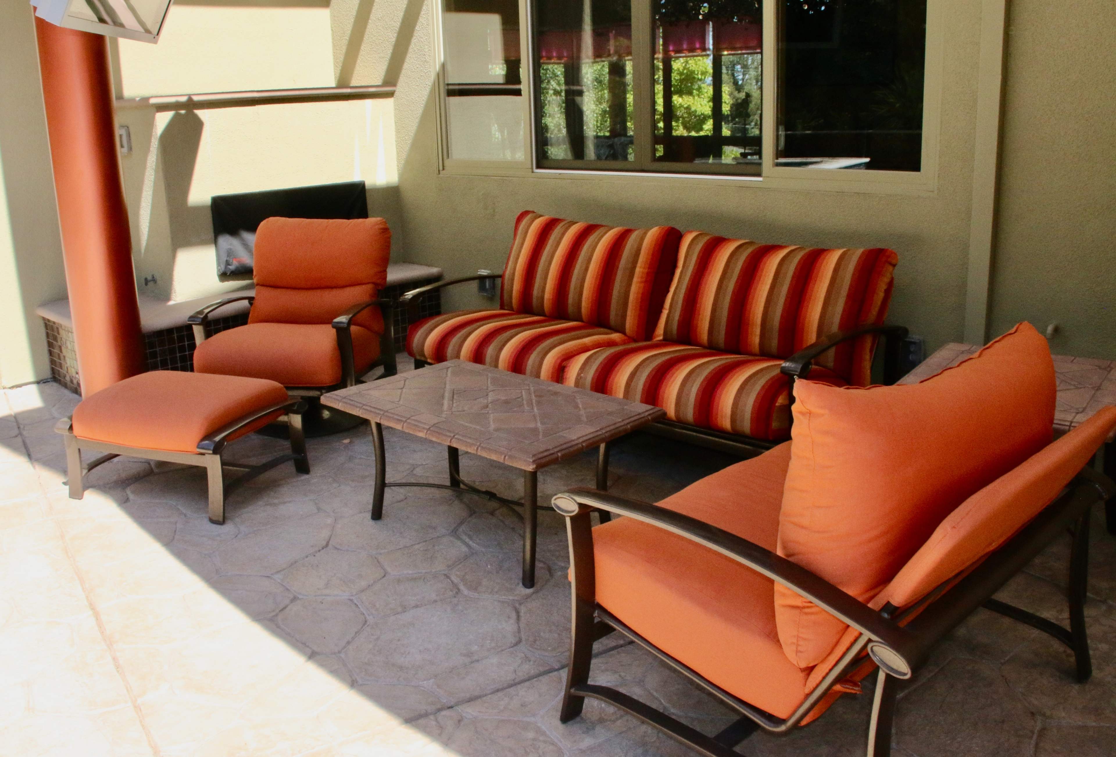 A patio seating area with a striped sofa, two orange chairs, and a coffee table on a textured stone floor.