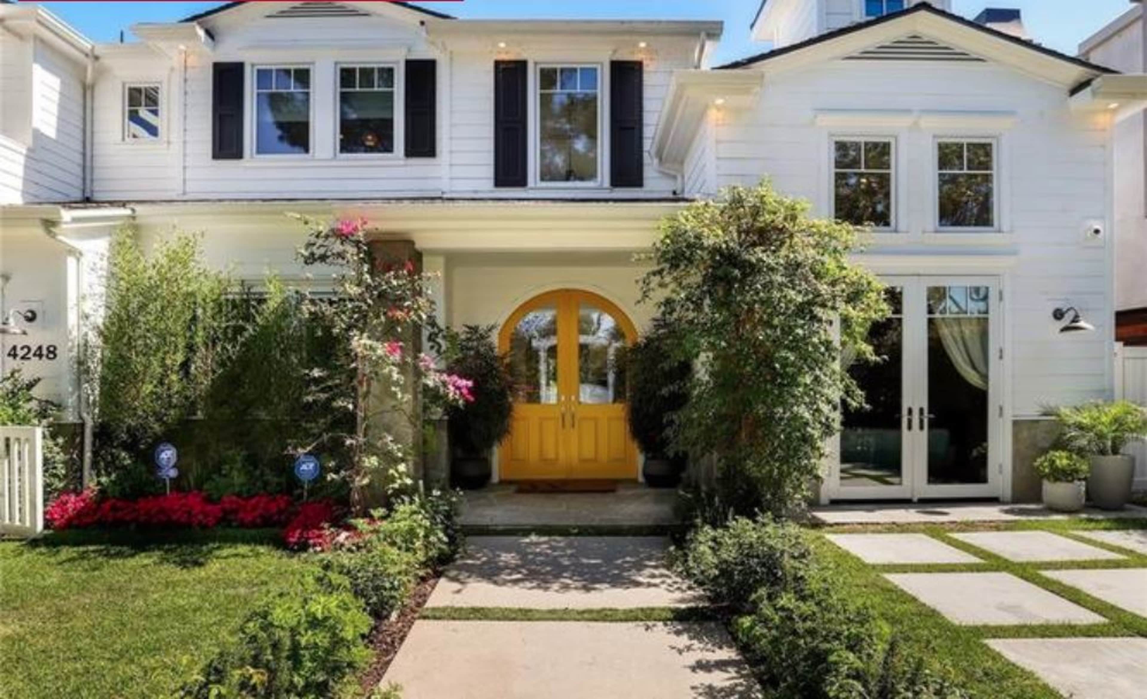 A white two-story house features a bright yellow front door, surrounded by landscaped gardens and a stone pathway.