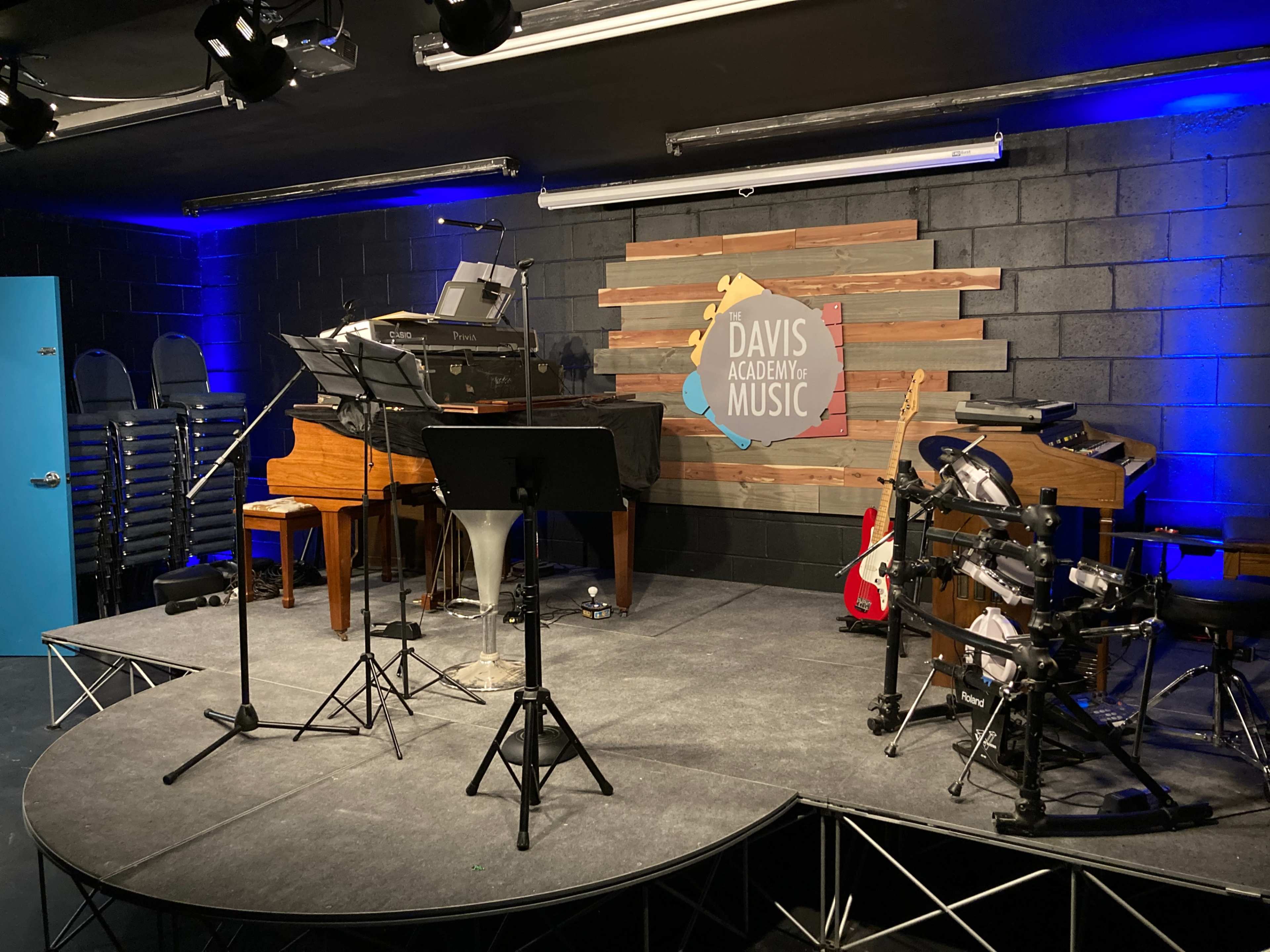 A music performance stage featuring a piano, a drum set, and various music stands, with a logo for Davis Academy of Music displayed on the back wall.