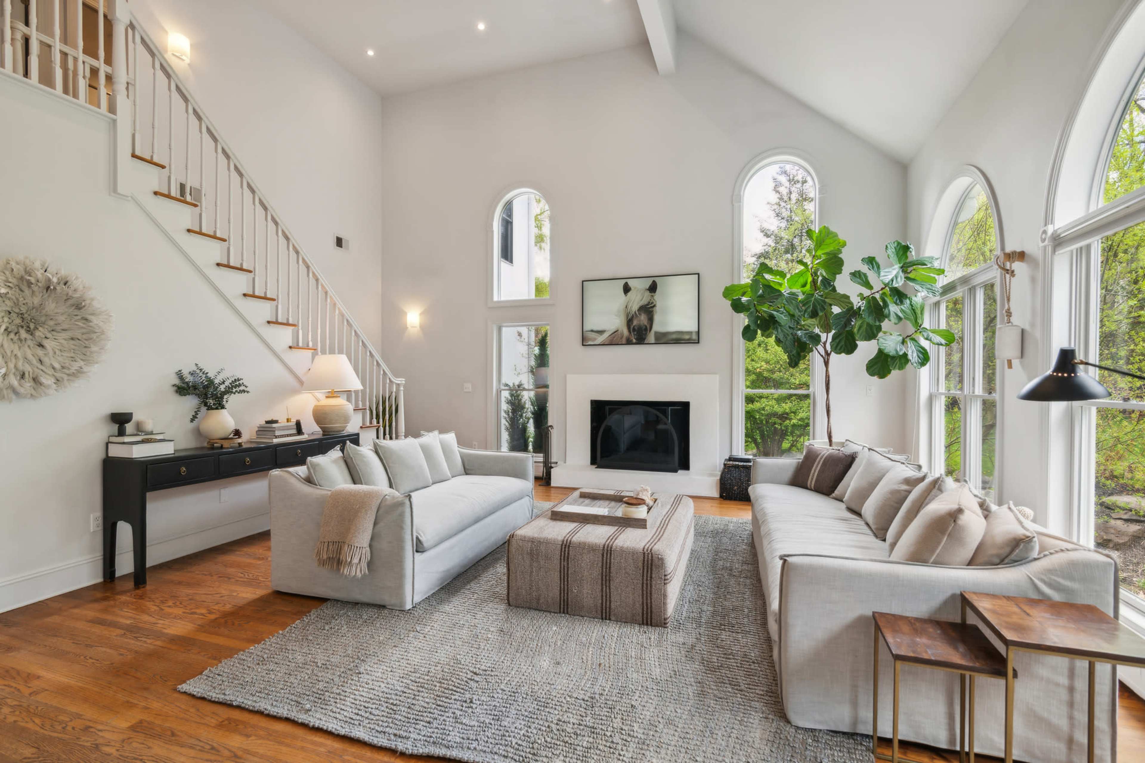 A modern living room with large windows, a beige sectional sofa, a coffee table, and a staircase leading to an upper level.