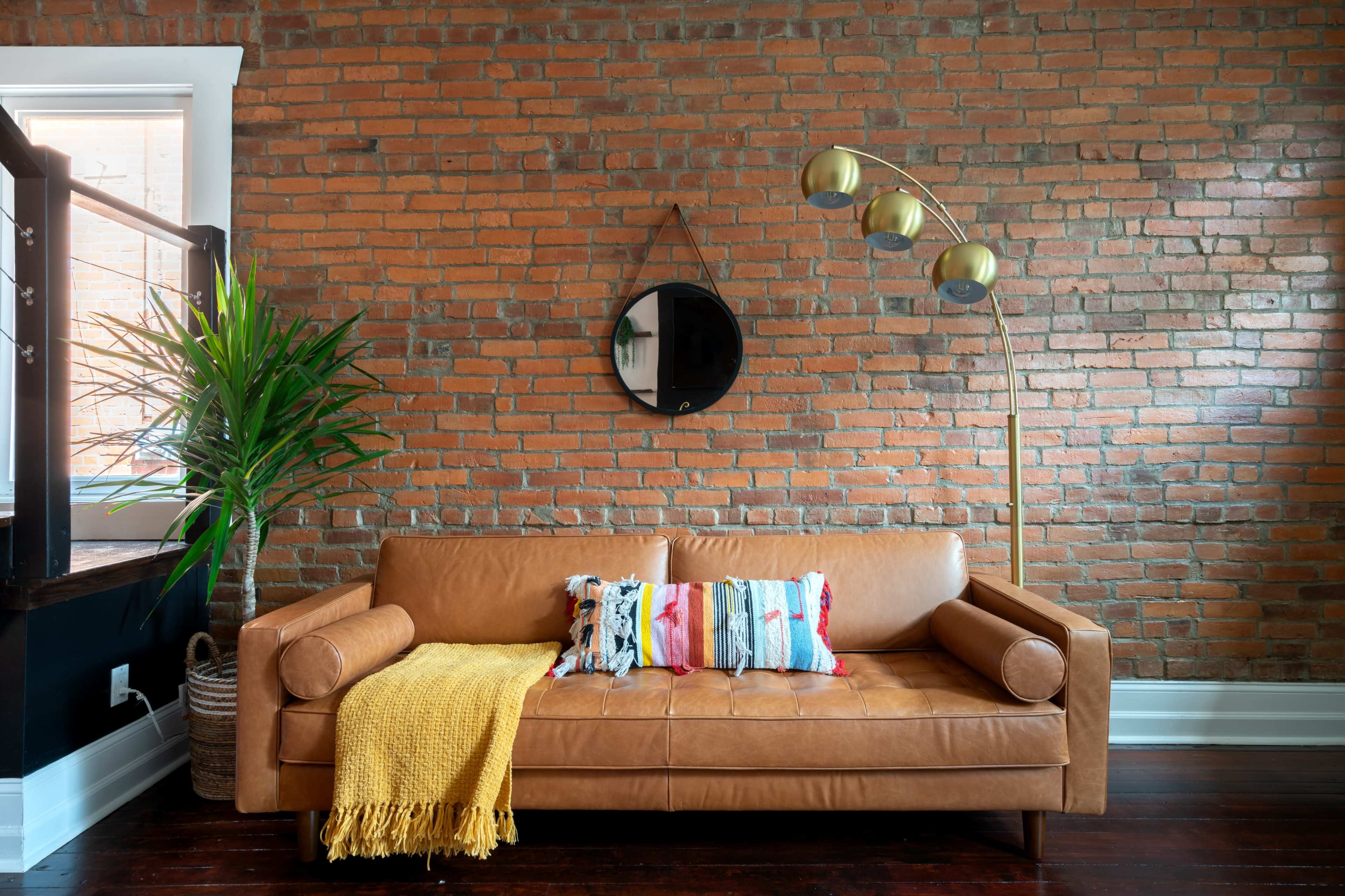 A tan leather sofa with colorful pillows and a yellow throw blanket is positioned against a brick wall, accompanied by a round mirror and a floor lamp.